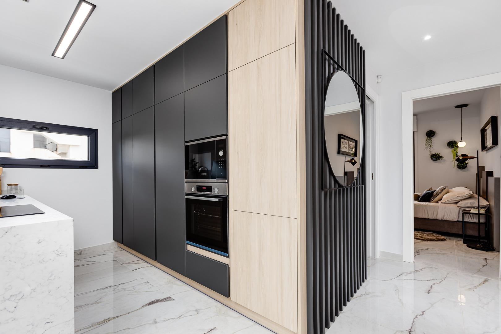 Modern kitchen with tall black cabinets, built-in microwave and oven, and light wood panels against a marble floor. The round mirror sits on a black slatted divider near a doorway to a bedroom.