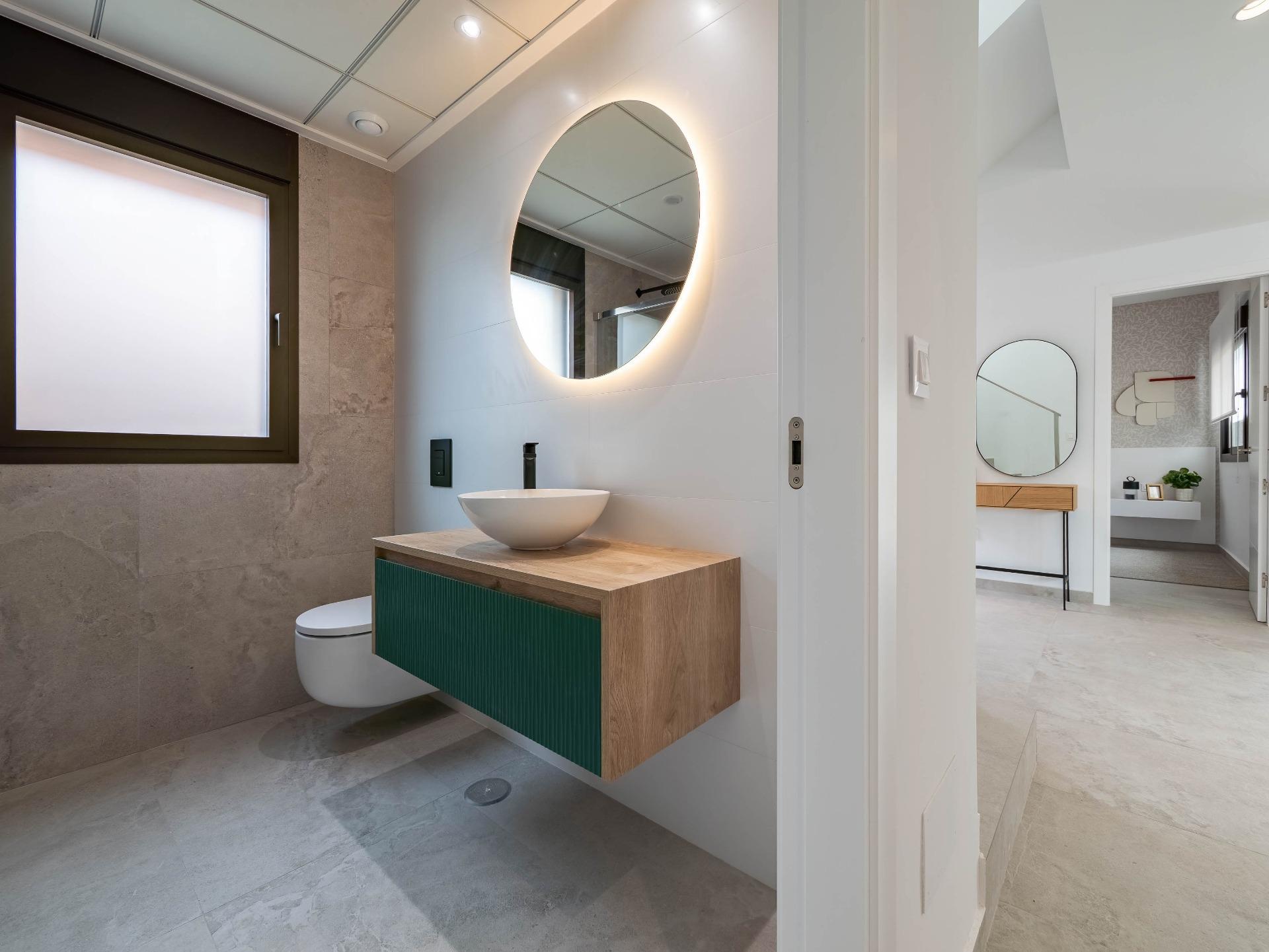 Contemporary bathroom with a round backlit mirror above a vessel sink on a wood-and-green vanity, gray tiled walls, and frosted window.