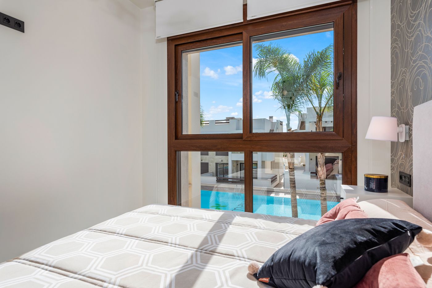 Bedroom with a large wooden-framed window overlooking a sunny pool area and palm trees; bed with geometric duvet and pink/black pillows nearby.