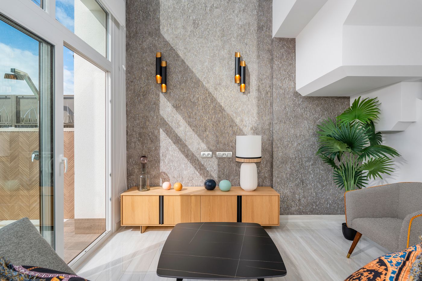 Bright modern living room with a wooden sideboard, decorative vases, and a textured wall under wall sconces by a large glass door.