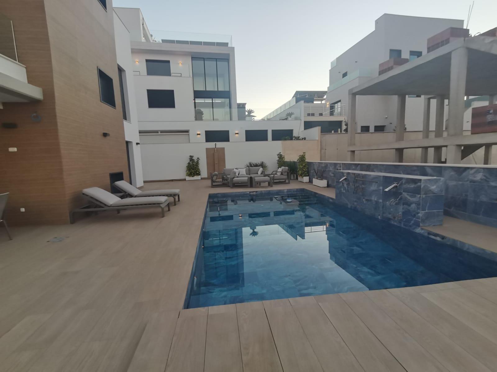 Modern villa courtyard with a rectangular blue-tiled pool, beige deck, and white contemporary buildings surrounding it; lounge chairs along the left edge.