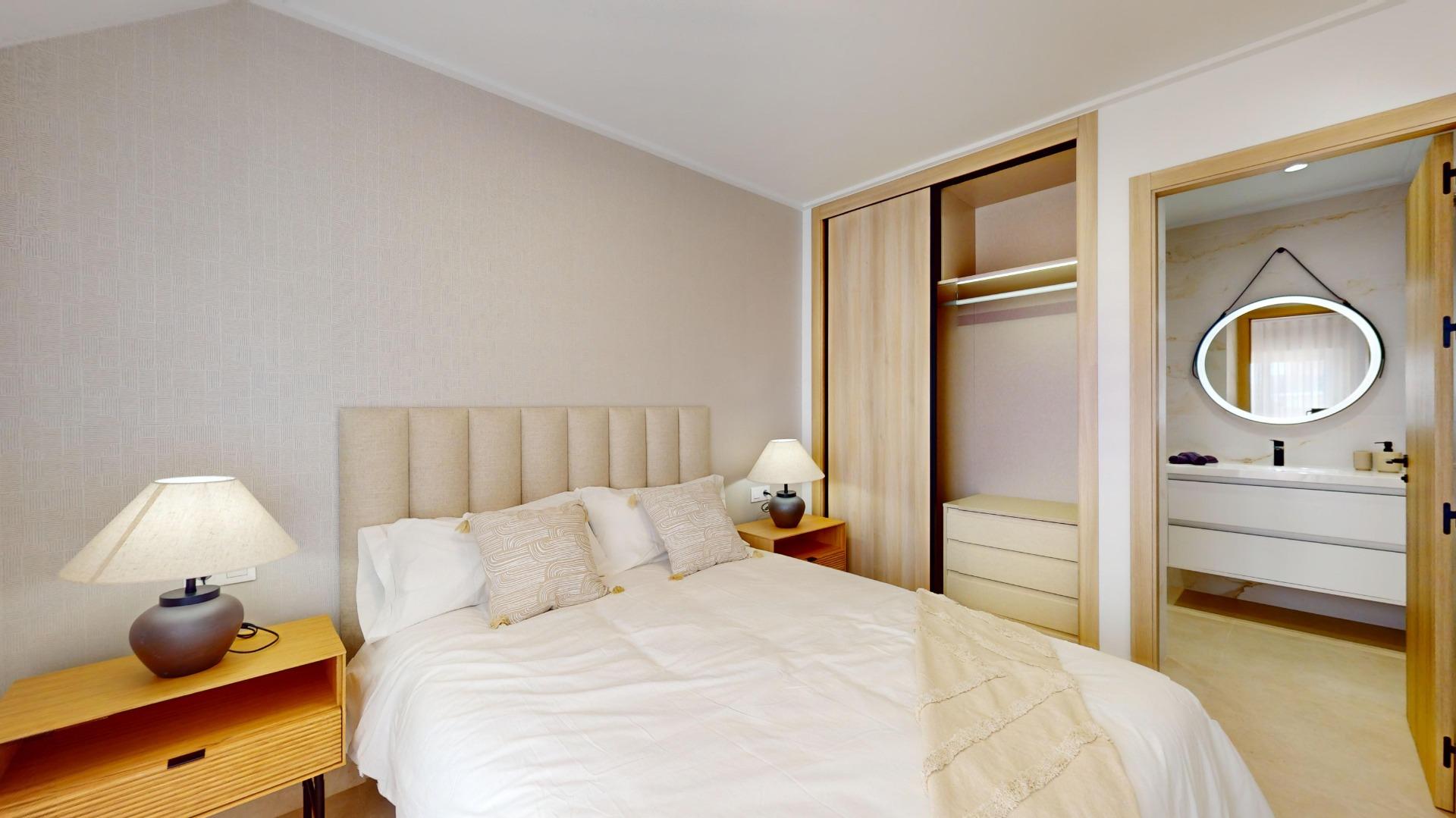 Neutral bedroom with padded headboard, white bedding, two bedside tables and lamps; open wardrobe and a bathroom with a round mirror visible through the door.