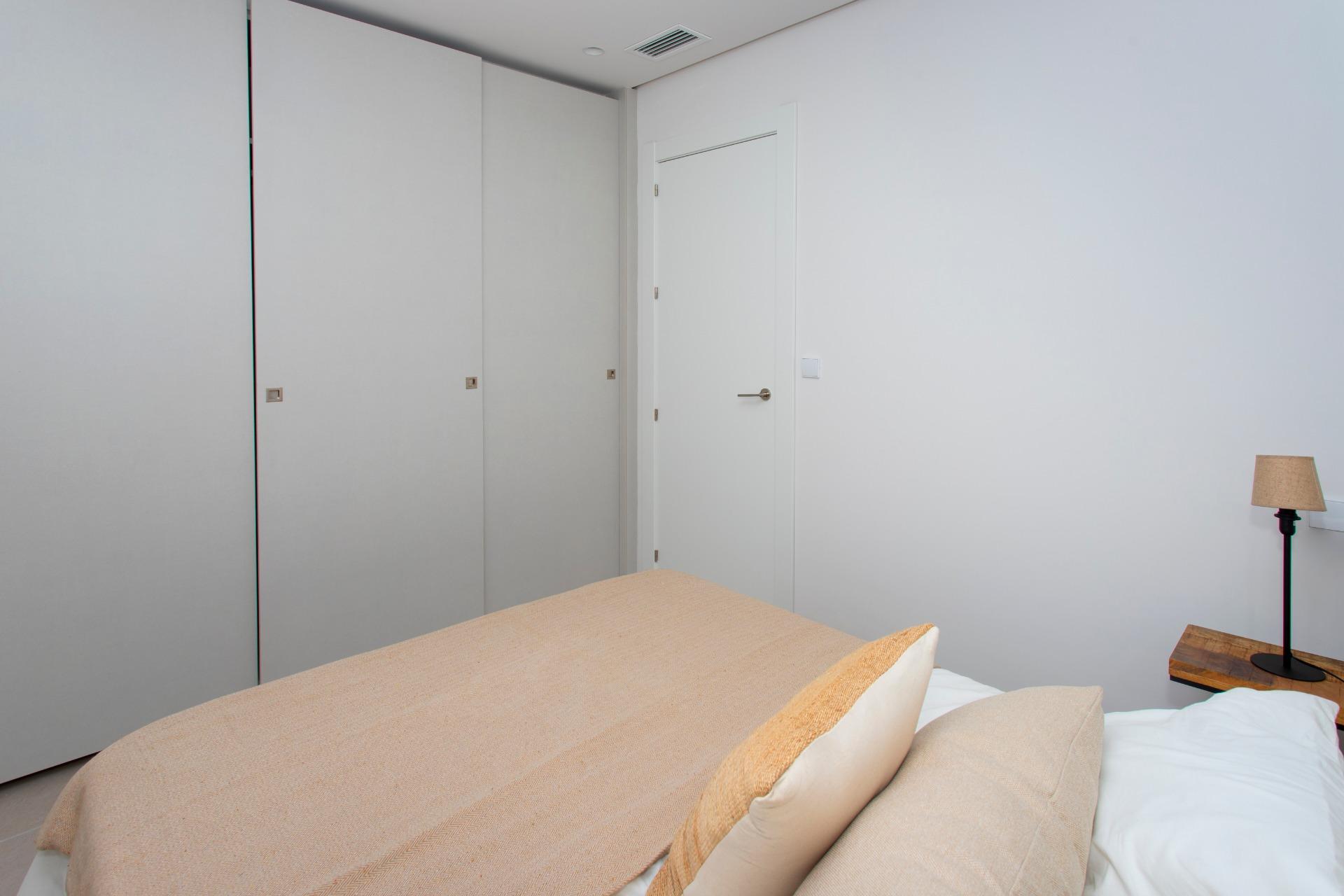 Neutral-toned bedroom featuring a bed with beige linens in the foreground, grey built-in wardrobes along the left wall, a white door on the far wall, and a small bedside lamp on a wooden table to the right.