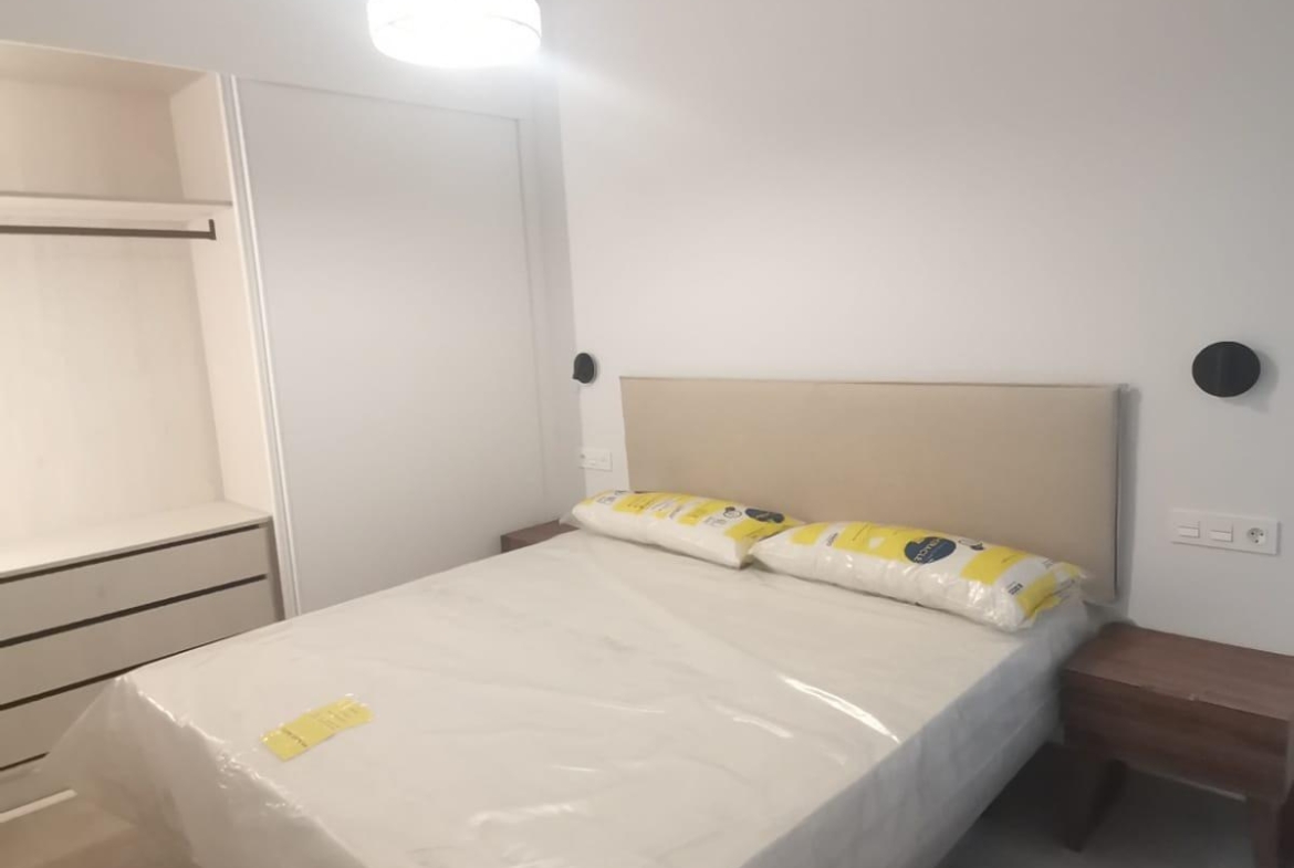 Bedroom with a bed wrapped in plastic, beige headboard, and two wooden nightstands on either side in a bright, modern room. An open wardrobe is visible on the left, and a ceiling pendant light hangs overhead.