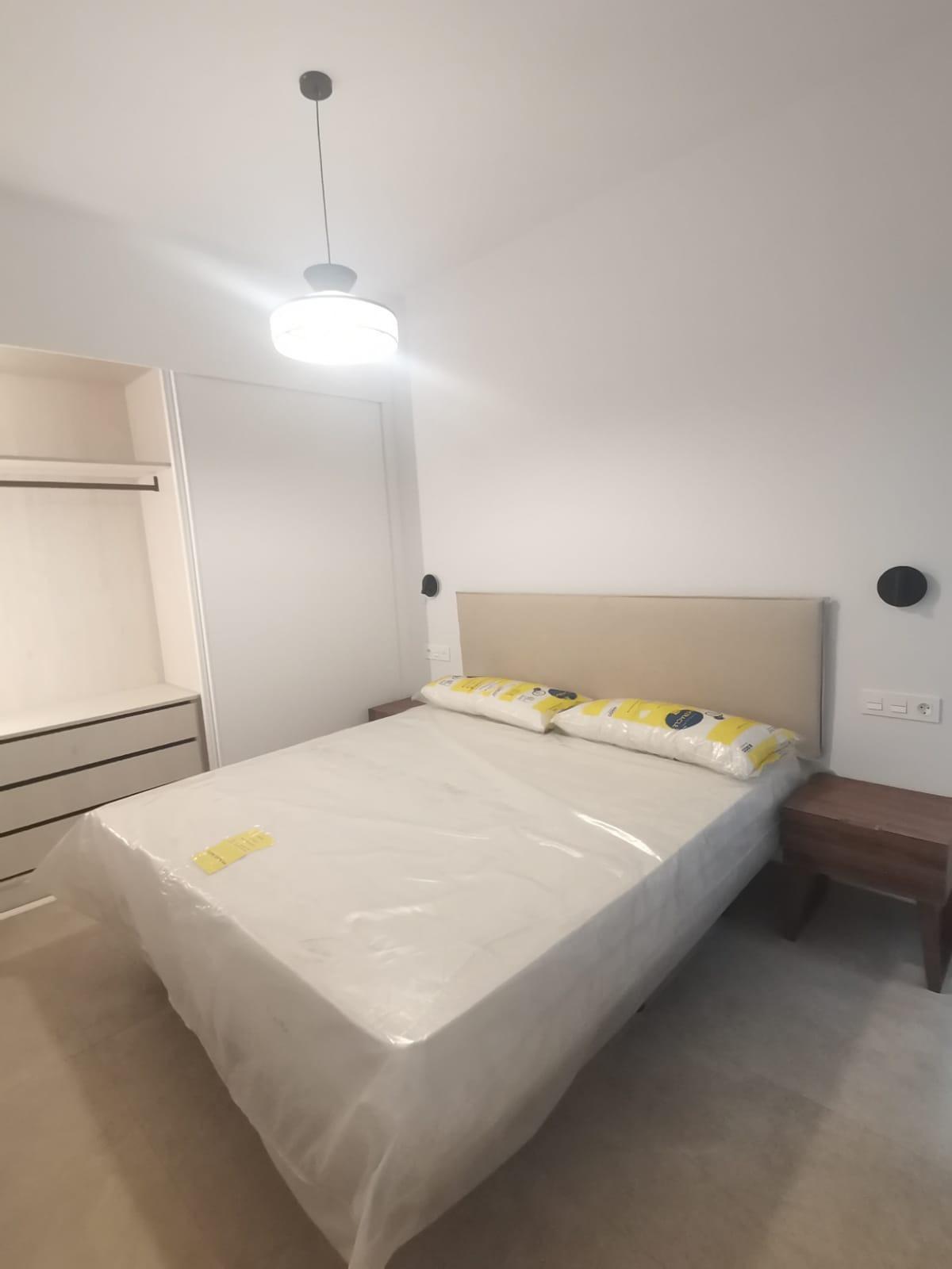 Bedroom with a bed wrapped in plastic, beige headboard, and two wooden nightstands on either side in a bright, modern room. An open wardrobe is visible on the left, and a ceiling pendant light hangs overhead.