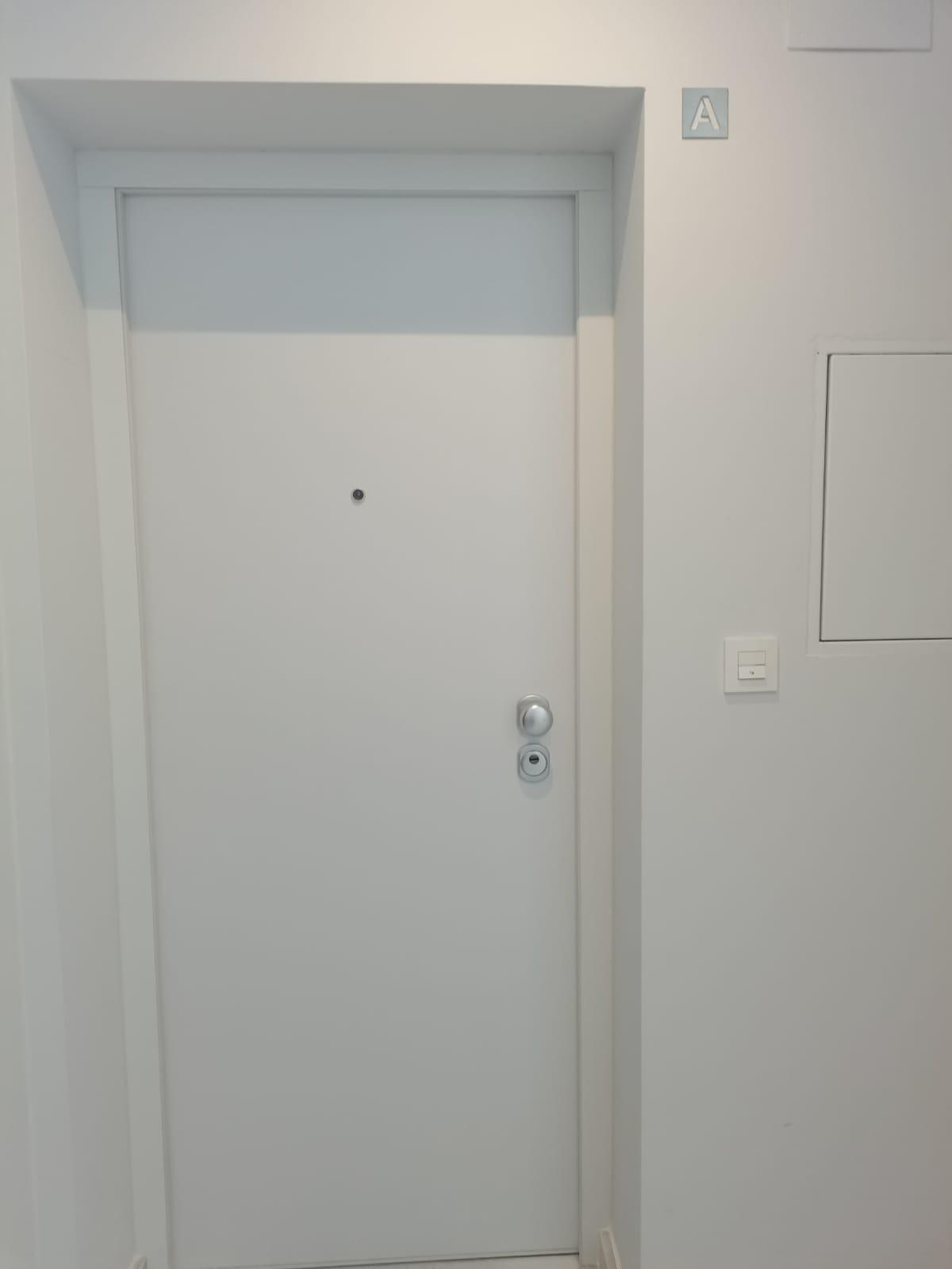 White interior door with a peephole and two locks in a clean hallway, with an 'A' sign on the wall to the right.