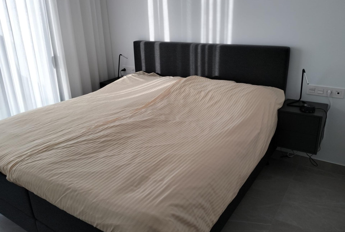 Bed with beige striped duvet and dark upholstered headboard flanked by two black nightstands and lamps on each side.