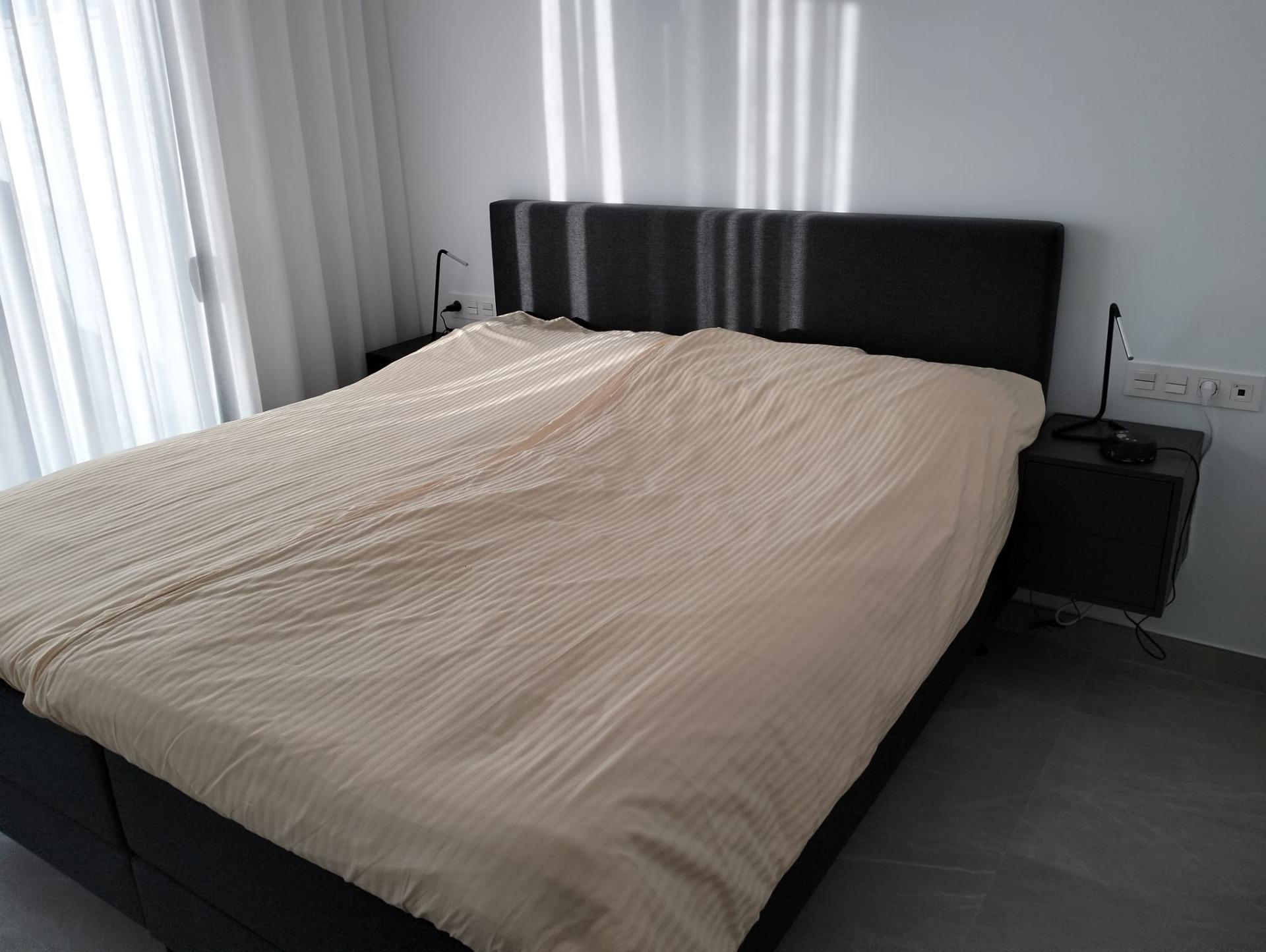 Bed with beige striped duvet and dark upholstered headboard flanked by two black nightstands and lamps on each side.