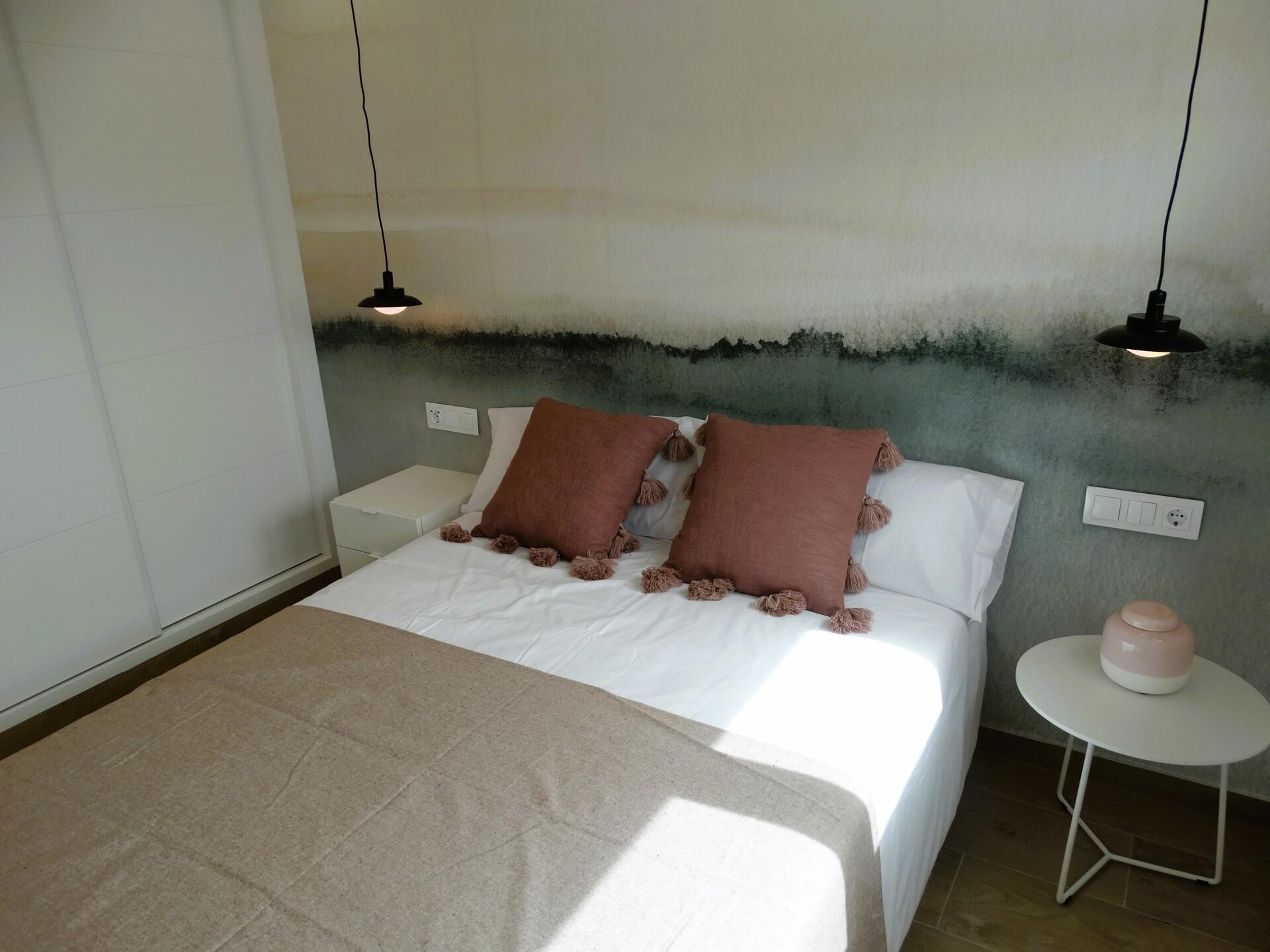 Modern bedroom with a white bed, taupe bed runner, and two rust-colored tassel pillows against a textured, two-tone wall; black pendant lights hang over each side of the bed.