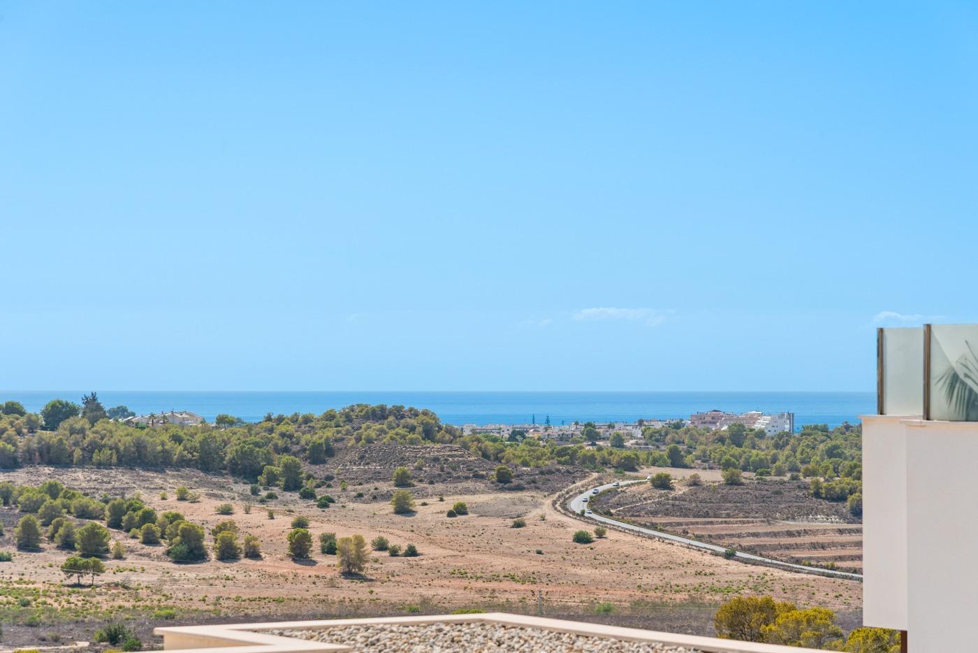 Coastal panorama with a bright blue sky, dry brown fields, scattered trees, and a winding road toward the distant sea on the horizon.