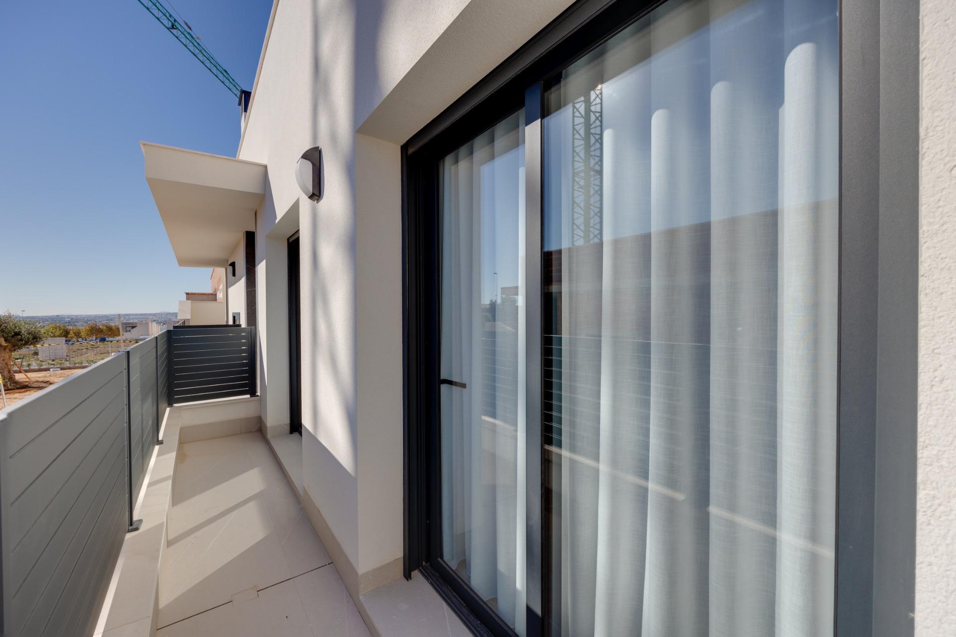 Balcony of a modern apartment with a large sliding glass door, sheer white curtains, and a gray railing under a blue sky.
