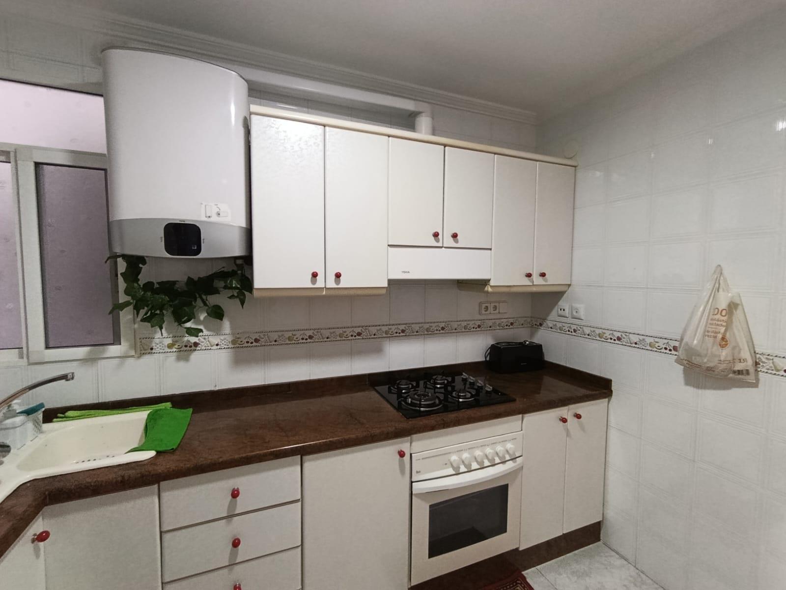 White kitchen with a brown countertop, wall-mounted cabinets, and a gas stovetop next to an oven. A boiler and a plant sit above the counter.