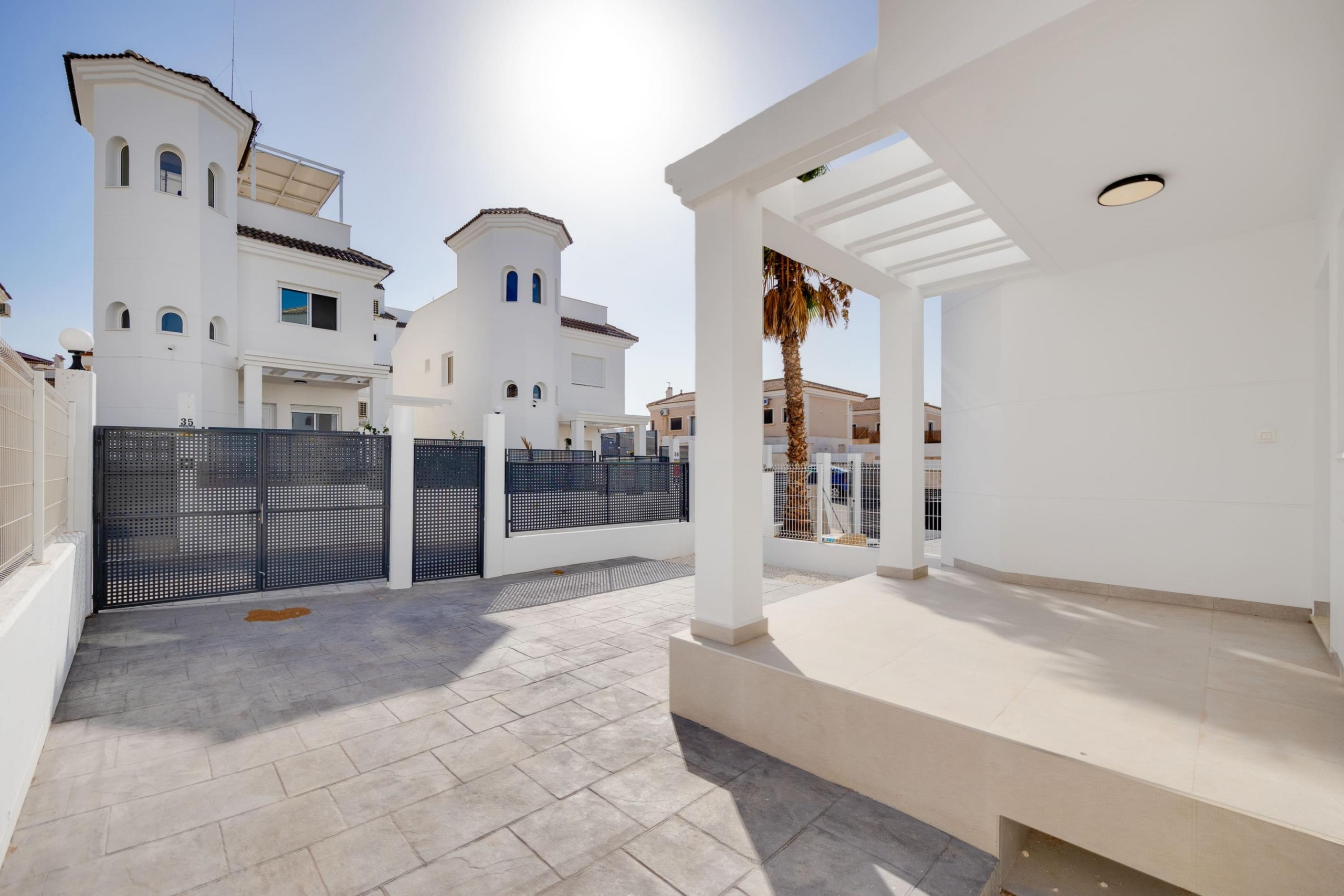 White modern villa complex with a black perforated gate, palm tree, and white arches under a clear sky.