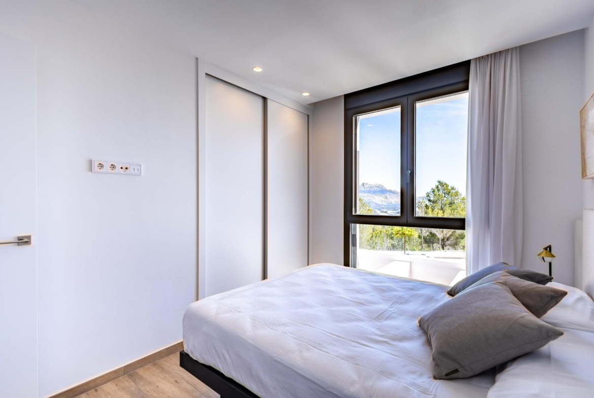 Modern bedroom with a large window showing mountains and trees, white walls, and a neatly made bed with gray pillows.