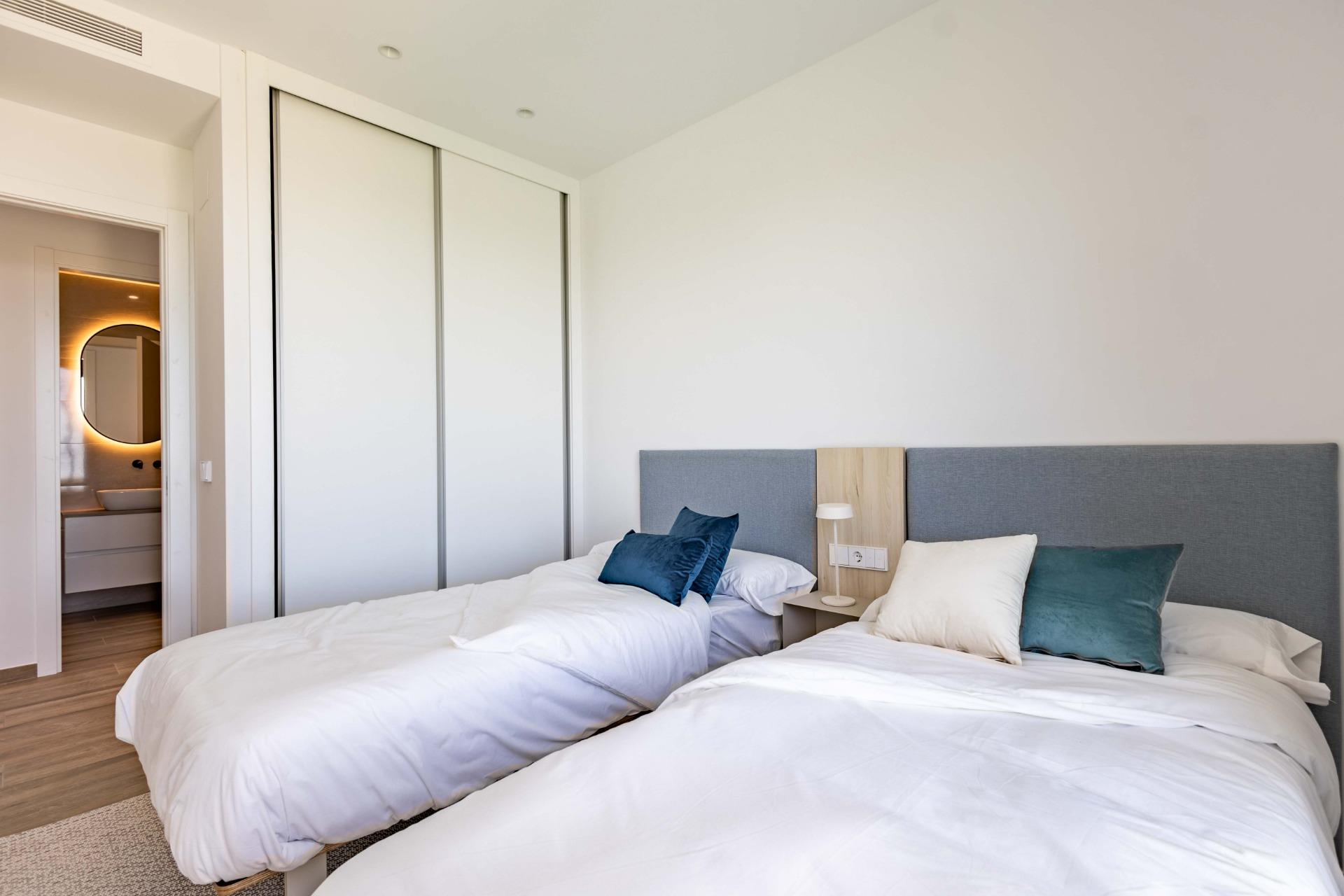 Twin beds with white duvets and blue/teal cushions in a modern hotel bedroom, with a large white sliding-door wardrobe and a bathroom doorway visible at left.