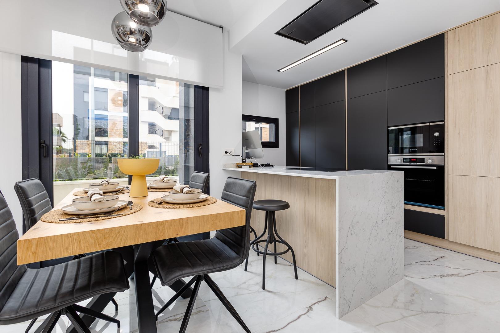 Modern kitchen and dining area with a wooden table set for six, black cabinet wall, marble island, and large windows.