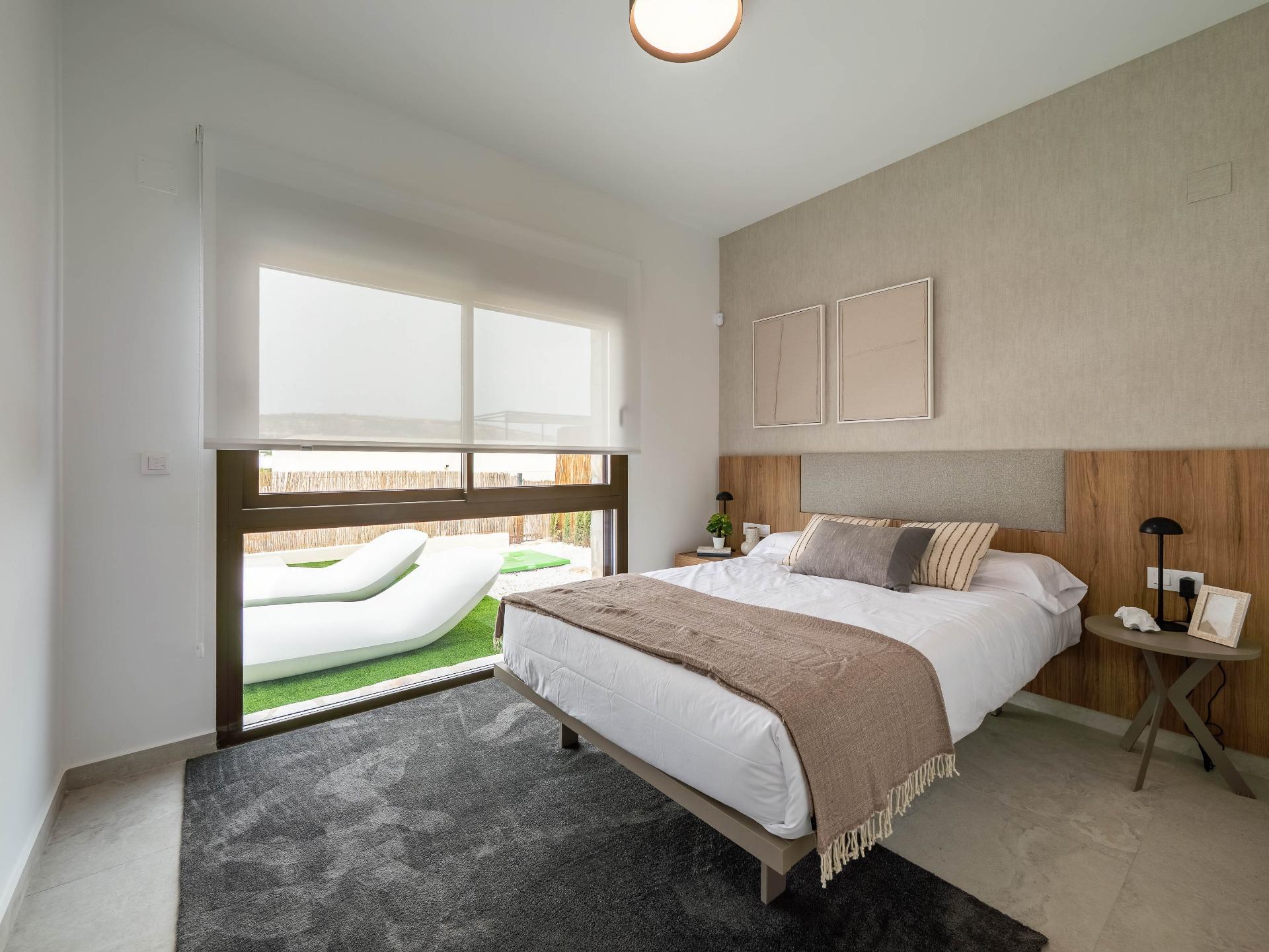 Modern bedroom with a low bed, beige headboard, two nightstands, and a glass door opening to a patio with white lounge chairs outside.