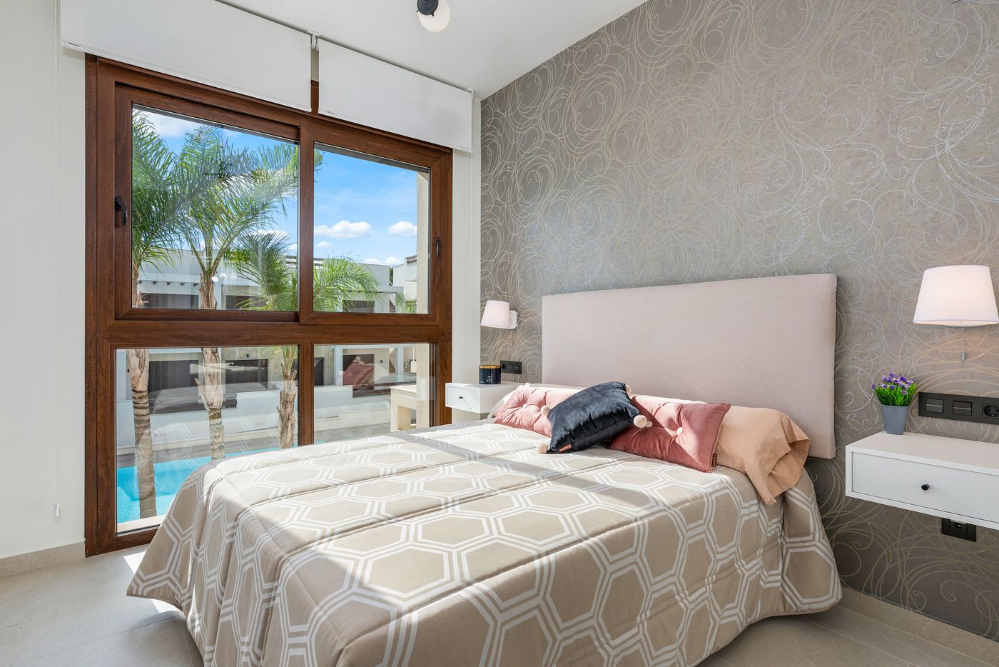 Bedroom with a large wood-framed window overlooking palm trees and a pool; bed has a beige geometric duvet and pink pillows to the right.