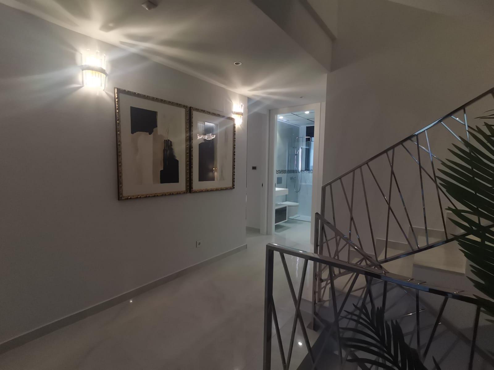 Hallway with two large framed abstract artworks, warm wall sconces, and a metal railing staircase on the right; door to a bright bathroom at the end.