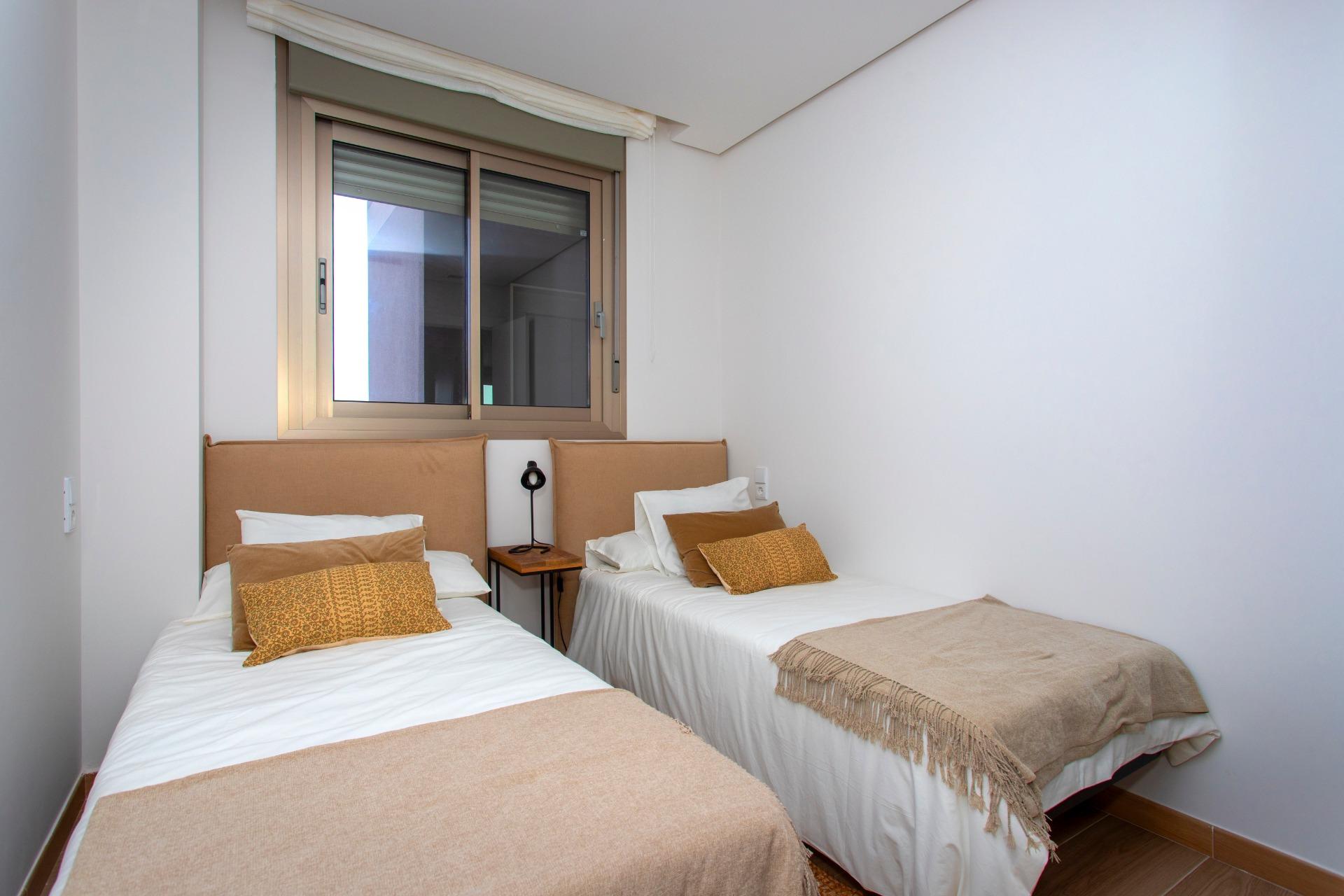Twin single beds with beige throws and decorative pillows in a bright hotel room, a small nightstand with a black lamp between them, and a window above.