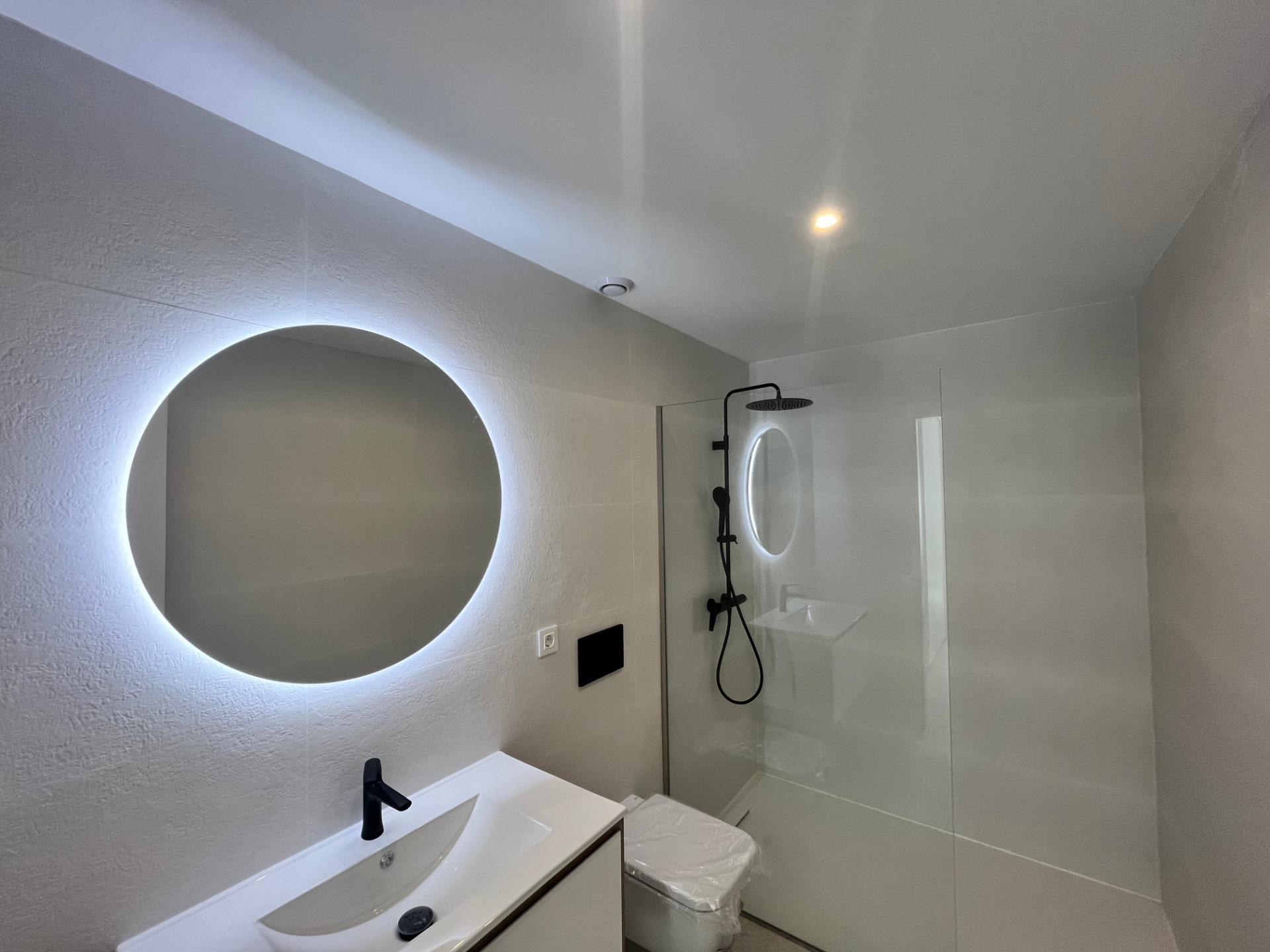 Modern bathroom with a round backlit mirror above a white vanity and a glass shower enclosure.