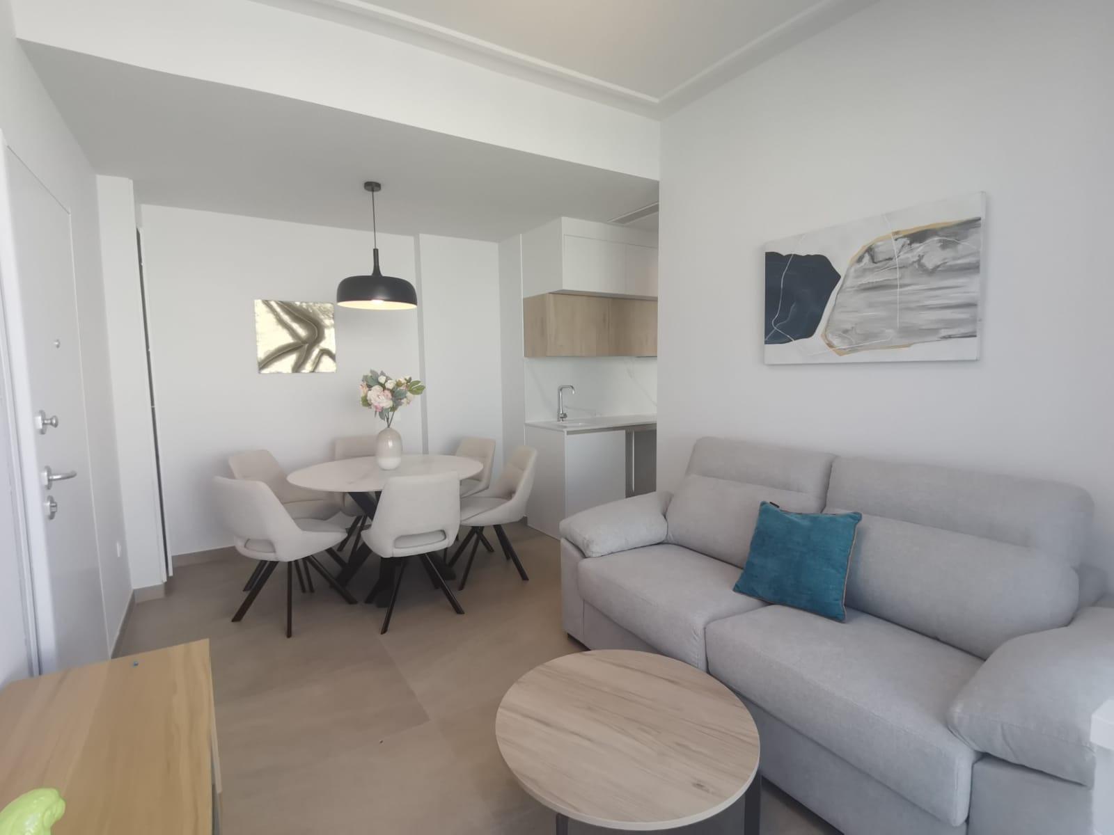 Open-plan living and dining area with a light gray sofa, blue cushion, round coffee tables, and a white dining table with six cream chairs; kitchenette in back and abstract wall art.