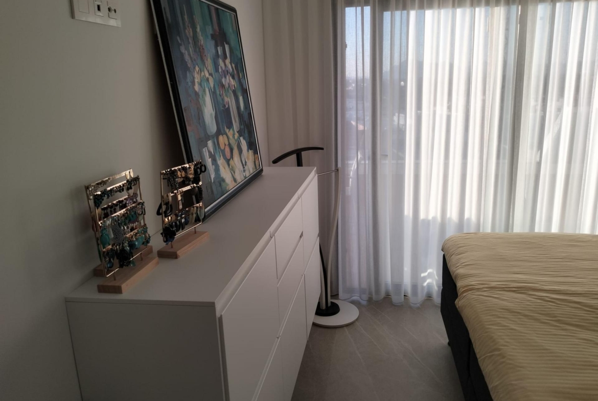 White dresser against a wall with a leaning colorful abstract painting and jewelry displays, next to sheer-curtained windows.