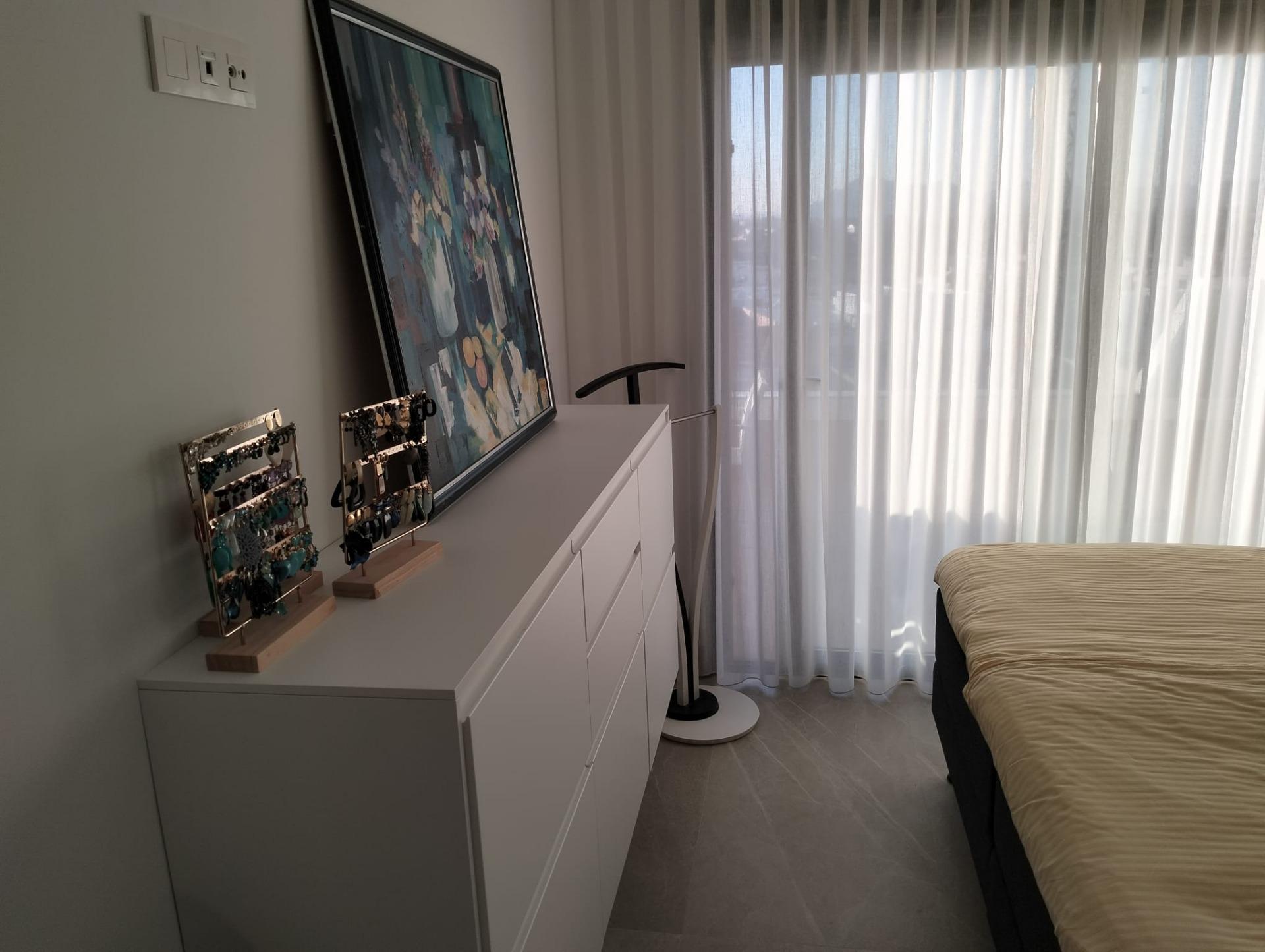 White dresser against a wall with a leaning colorful abstract painting and jewelry displays, next to sheer-curtained windows.
