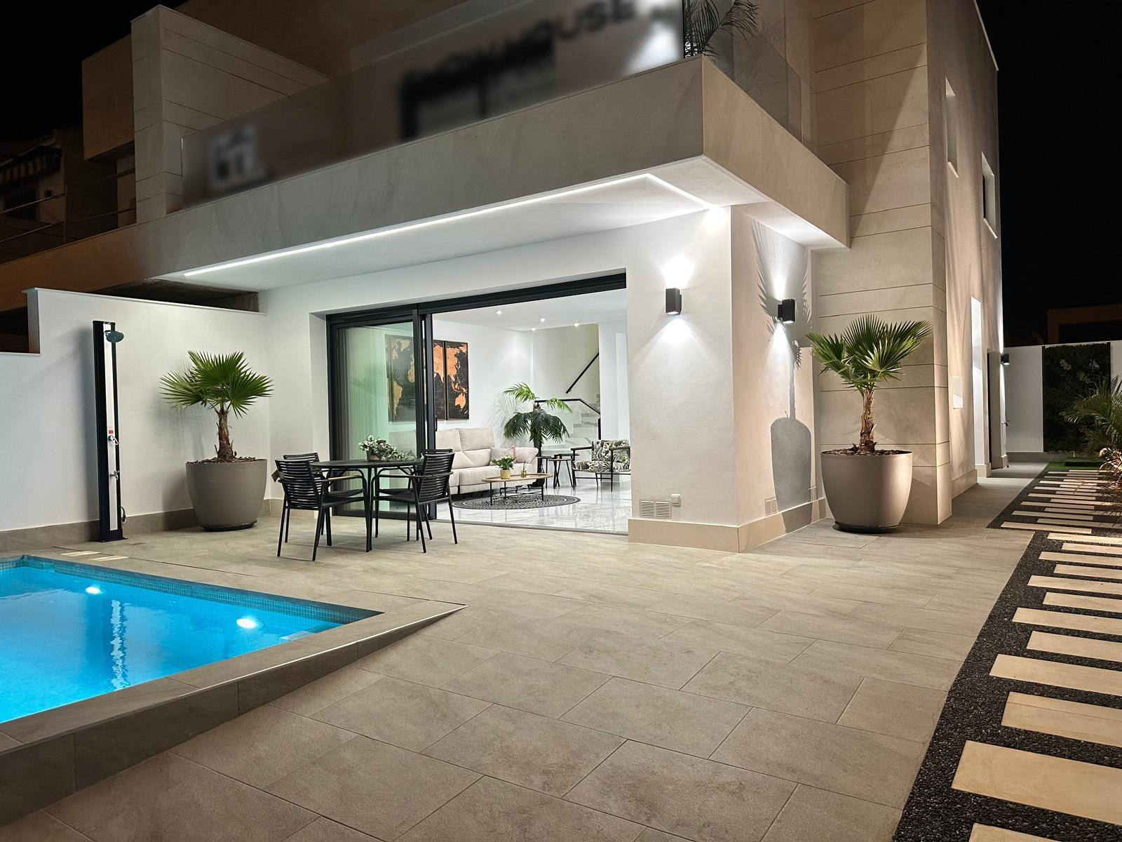 Night view of a modern white house with an illuminated patio, glass sliding doors opening to a seating area, and a rectangular pool; potted palms nearby.