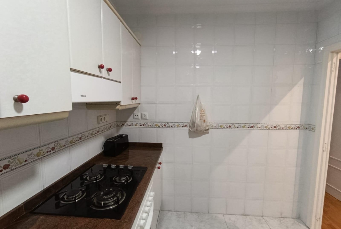 Compact white kitchen with glossy cabinets, red knobs, and a dark brown countertop gas hob. The tiled wall has a decorative border, and a toaster sits on the counter without clutter.