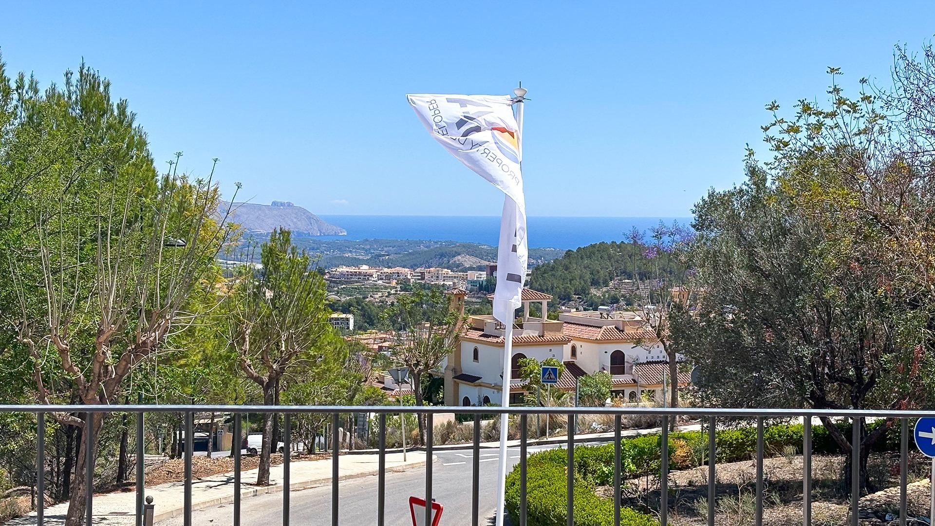 Coastal hillside town with red-tiled roofs, greenery, and a white flag fluttering on a pole in the foreground against a blue sea sky.