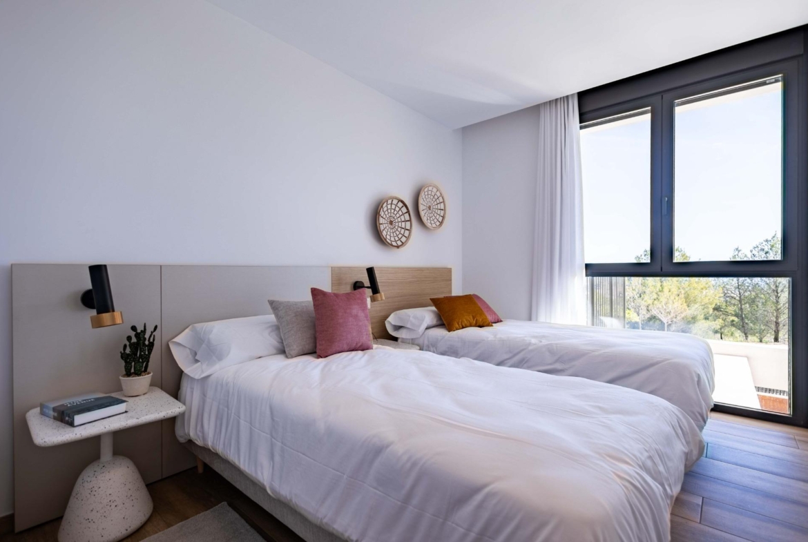 Twin beds in a bright hotel room with white linens, colorful pillows, and a large window letting in daylight.