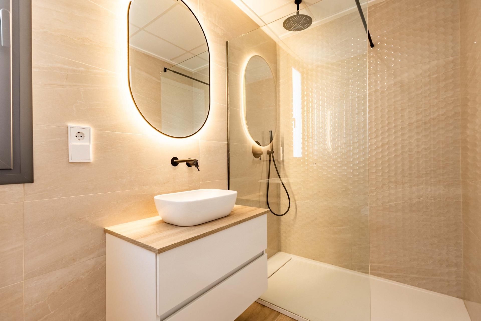 Modern bathroom interior with round backlit mirror above a white vessel sink on a wooden vanity and beige tile walls.