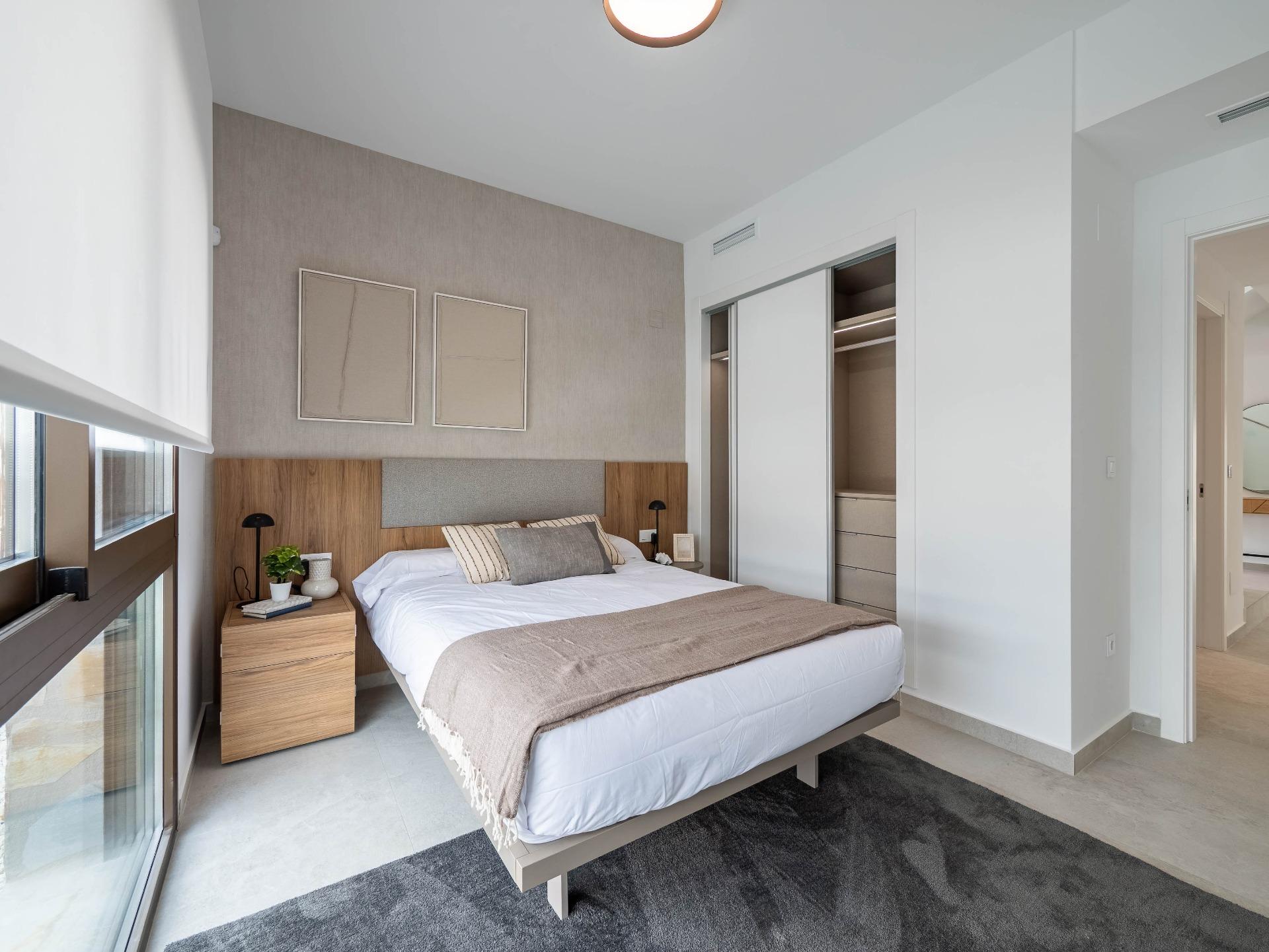 Modern hotel-style bedroom with a low platform bed, wooden headboard, beige bedding, and two matching nightstands near a large window sliding door. A built-in wardrobe with sliding doors is visible on the right. Neutral color palette and soft lighting create a calm retreat.