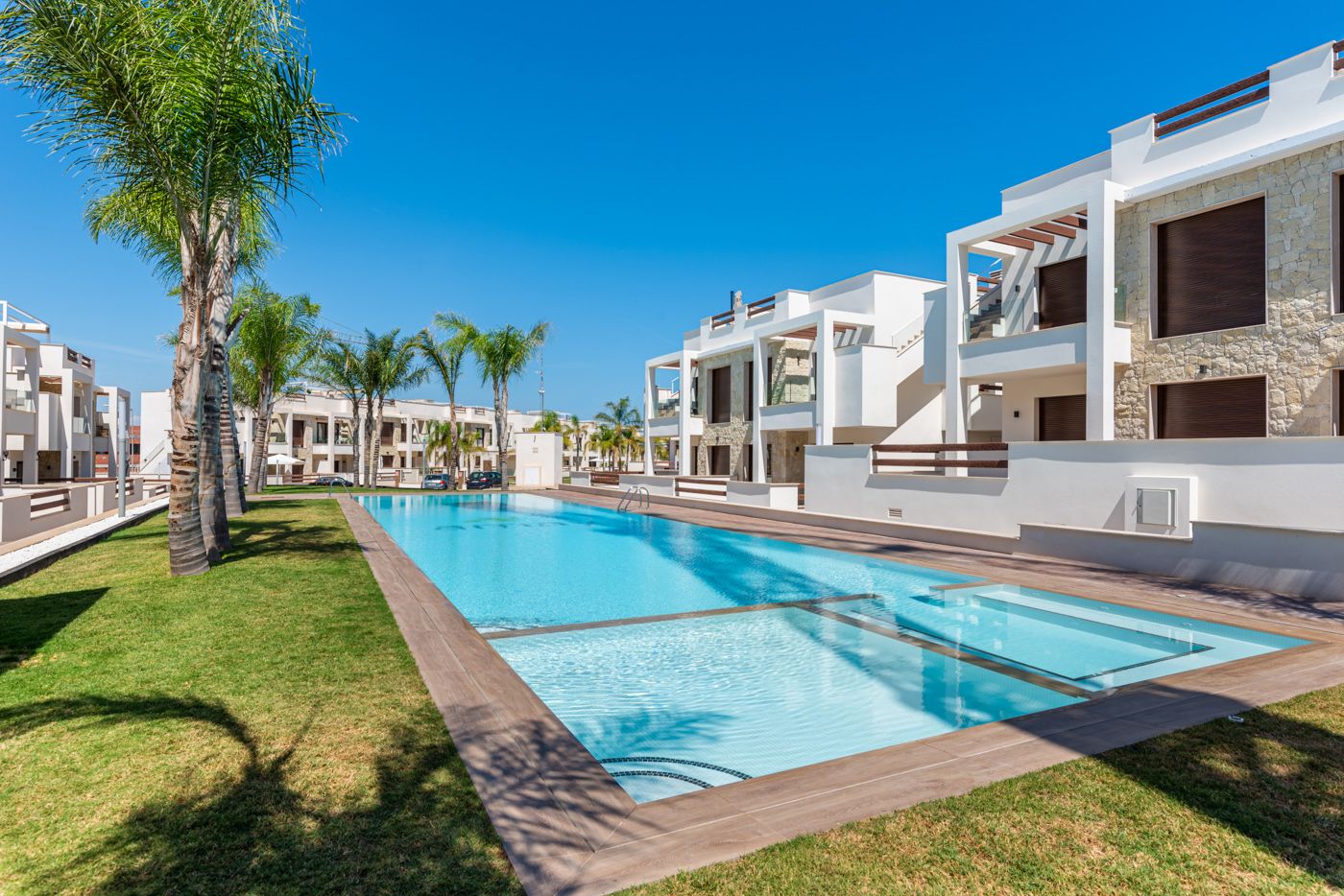 Resort-style courtyard with a long rectangular pool, palm trees, and white modern buildings under a clear blue sky.
