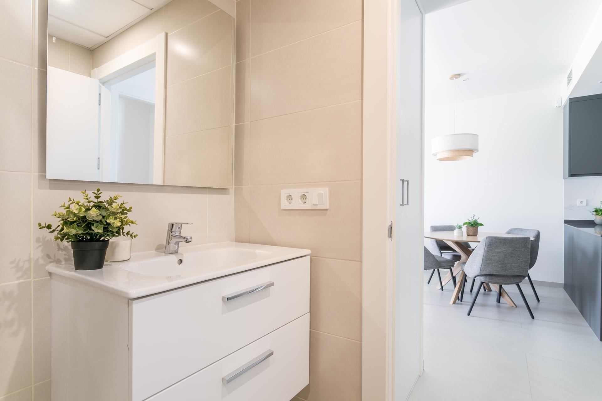Modern bathroom vanity with white drawers, ceramic sink, chrome faucet, and potted plant, opening to a bright dining area with a round table and grey chairs.