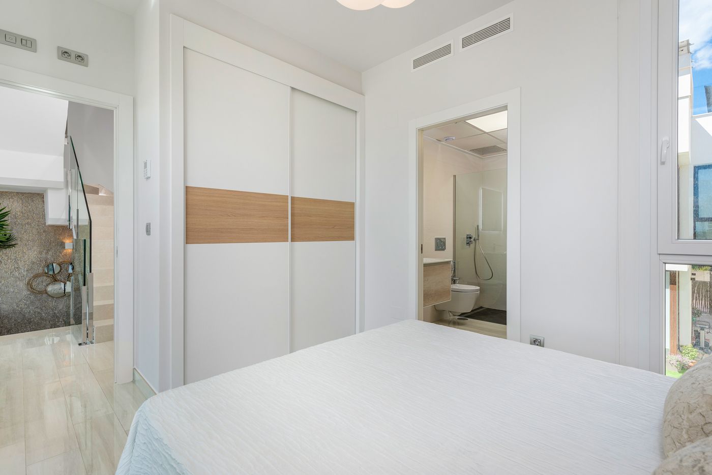 Bright modern bedroom with white walls, a double sliding wardrobe with a wood accent, and an open bathroom doorway opposite the bed.