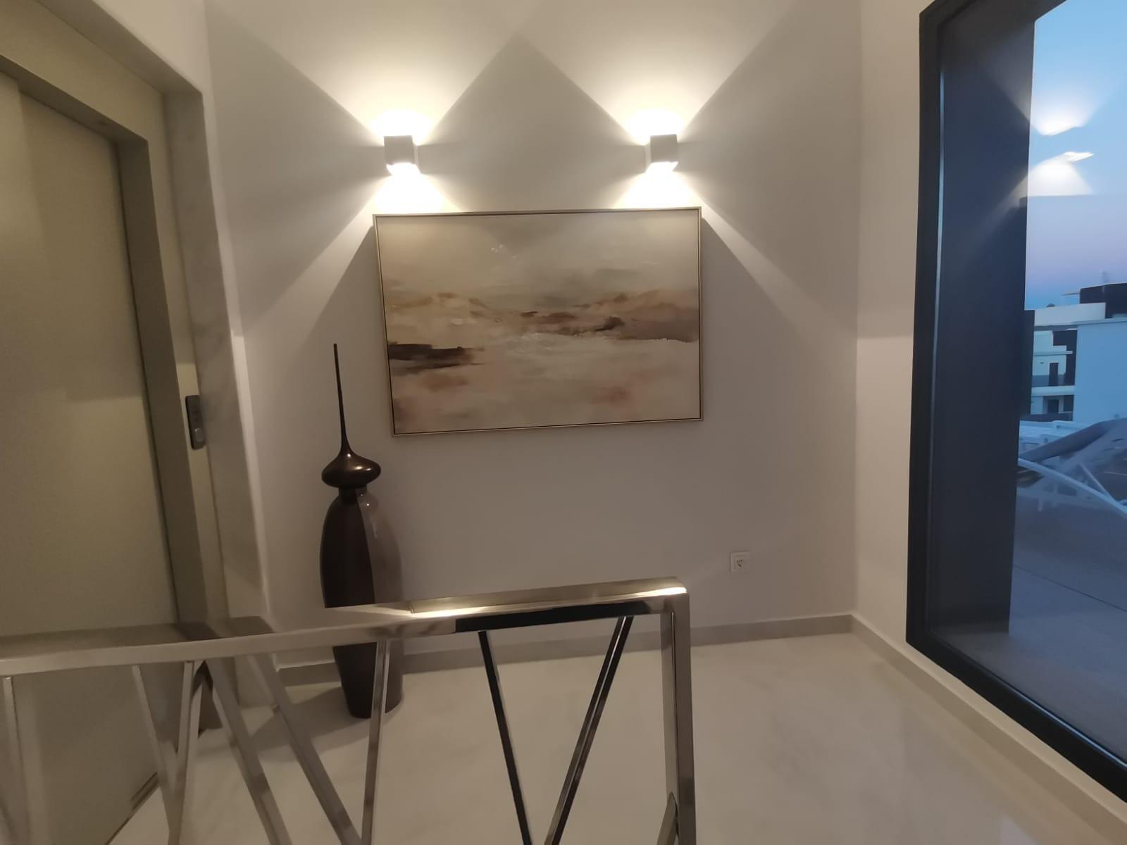 Modern hallway with abstract landscape painting, two wall sconces, a tall dark sculpture, glass railing, and a large window showing an evening city view.