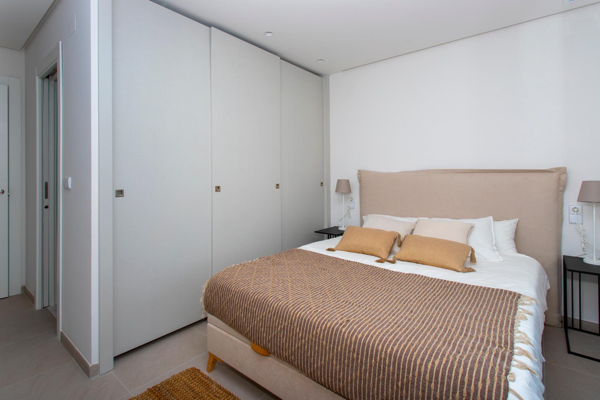 Bedroom with a long gray wardrobe on the left, beige upholstered bed, brown striped bedspread, and matching nightstands with lamps.