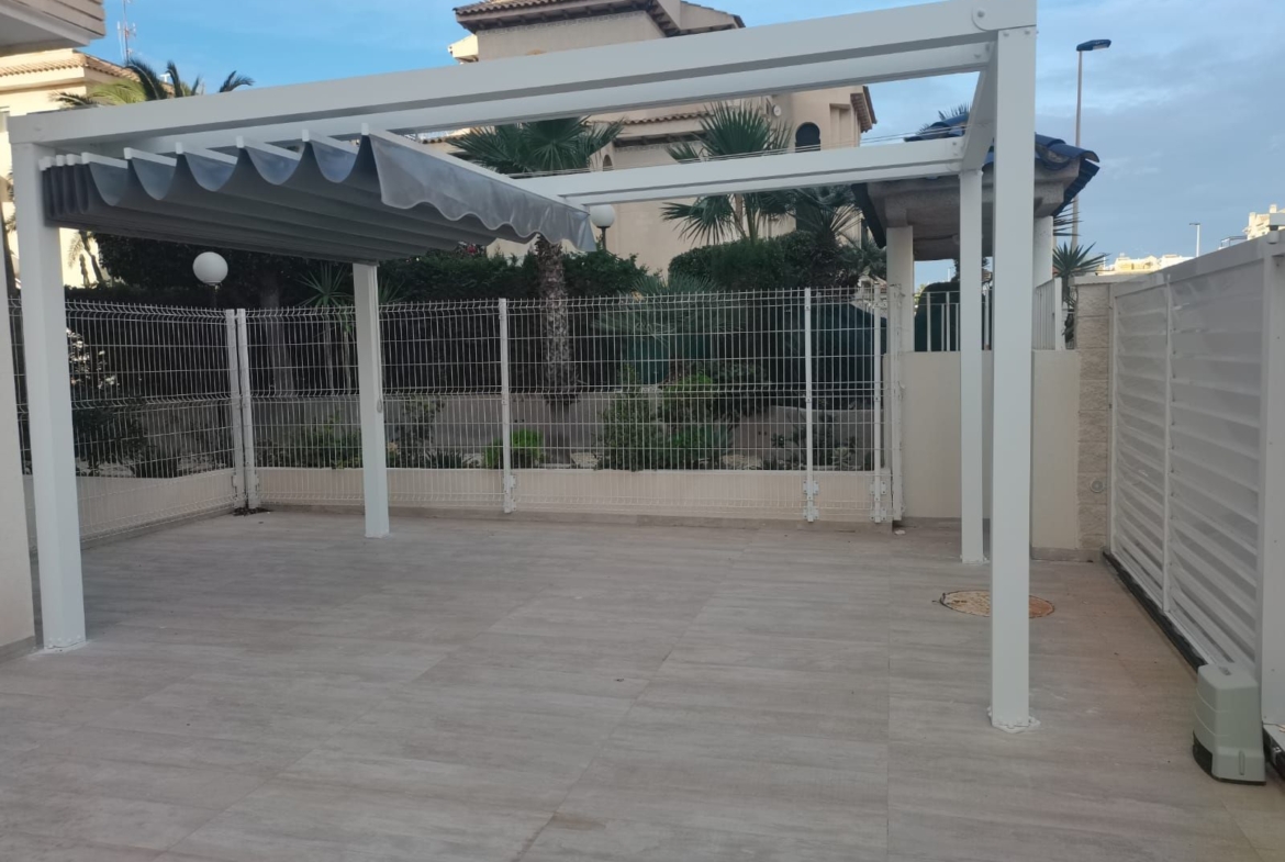 White metal pergola over a tiled patio with a gray retractable awning, fenced yard, and palm-treed houses in the background.