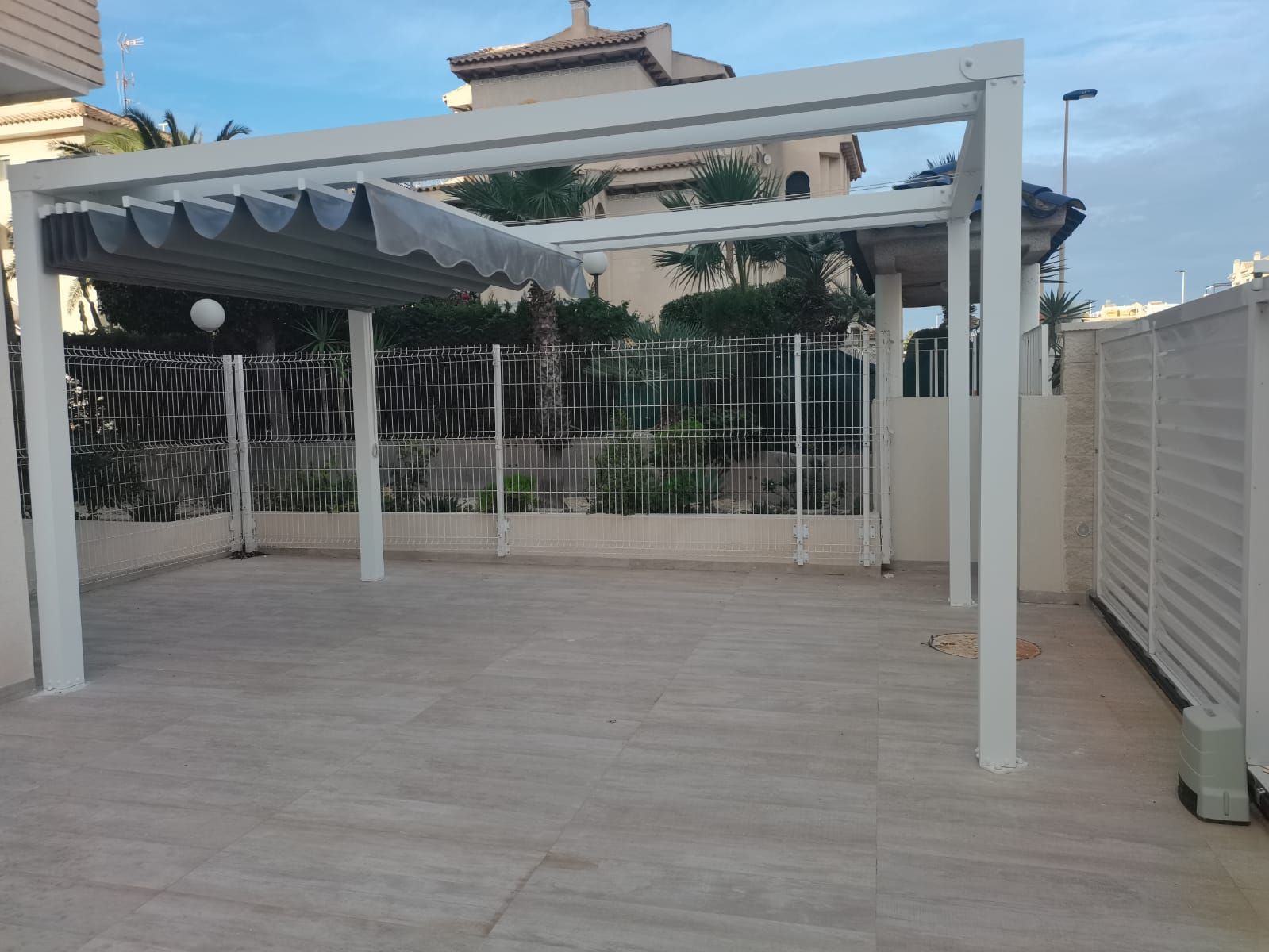 White metal pergola over a tiled patio with a gray retractable awning, fenced yard, and palm-treed houses in the background.