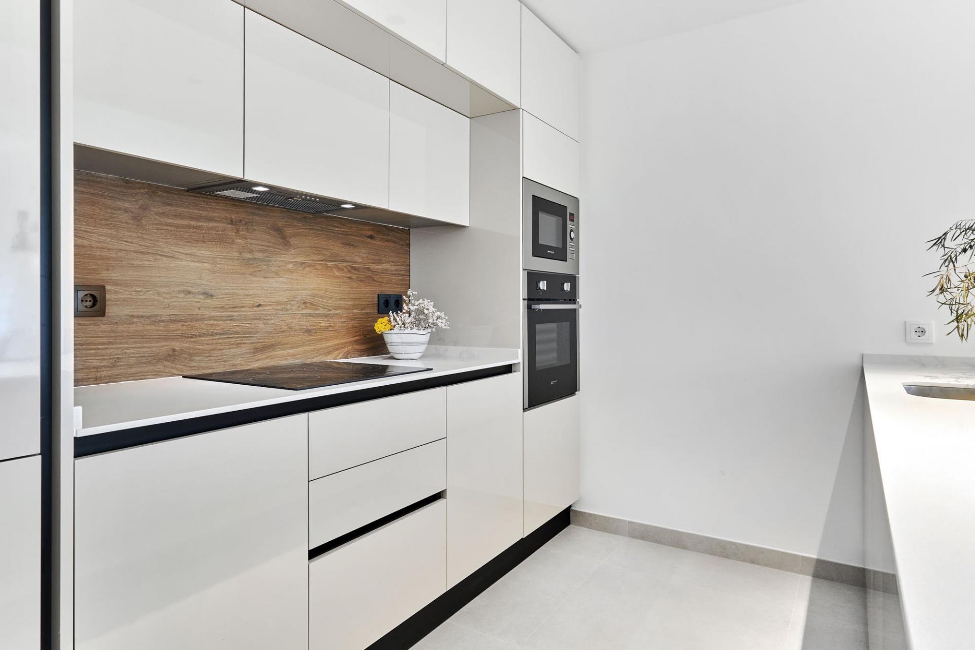 Minimalist white kitchen with a warm wood backsplash, built-in microwave and oven, and sleek handleless cabinetry.