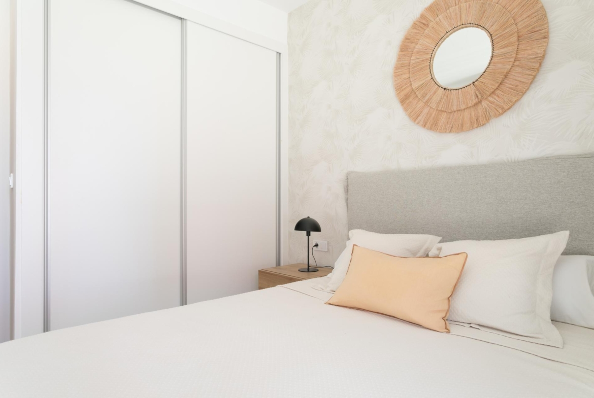 Cozy modern bedroom with white bedding, peach pillows, and a round woven mirror above a gray headboard.