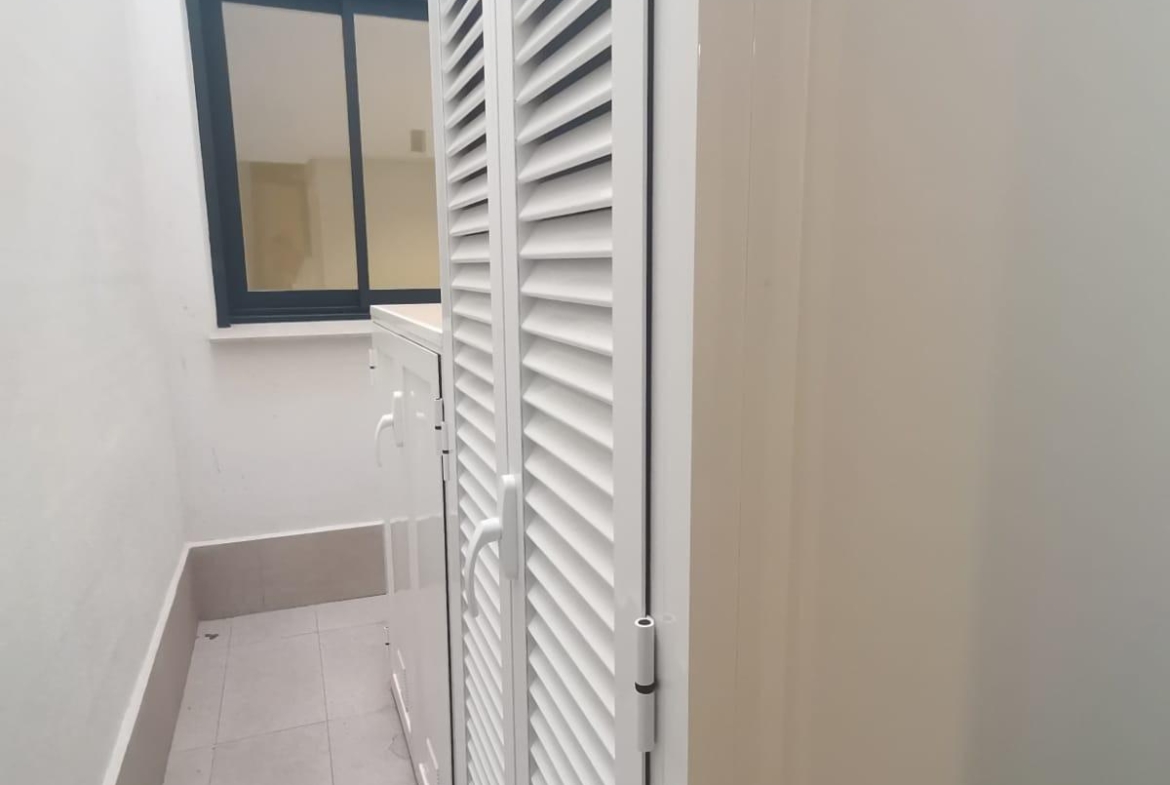 Narrow outdoor corridor with white louvered doors along one side, a window at the far end, and light gray tiled flooring.