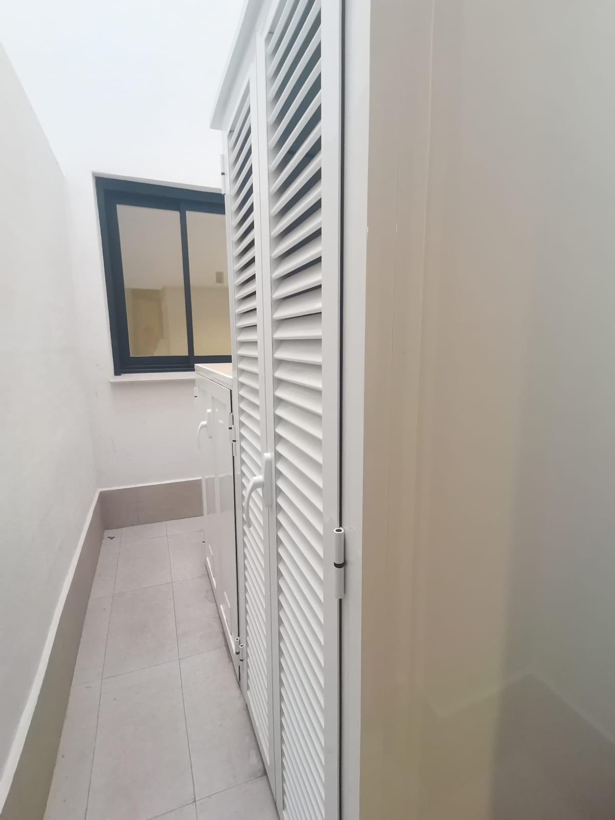 Narrow outdoor corridor with white louvered doors along one side, a window at the far end, and light gray tiled flooring.