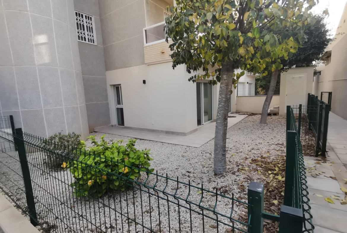 Backyard area of a modern building with light gray walls, sliding glass door, two trees, and a gravel yard enclosed by a green metal fence.