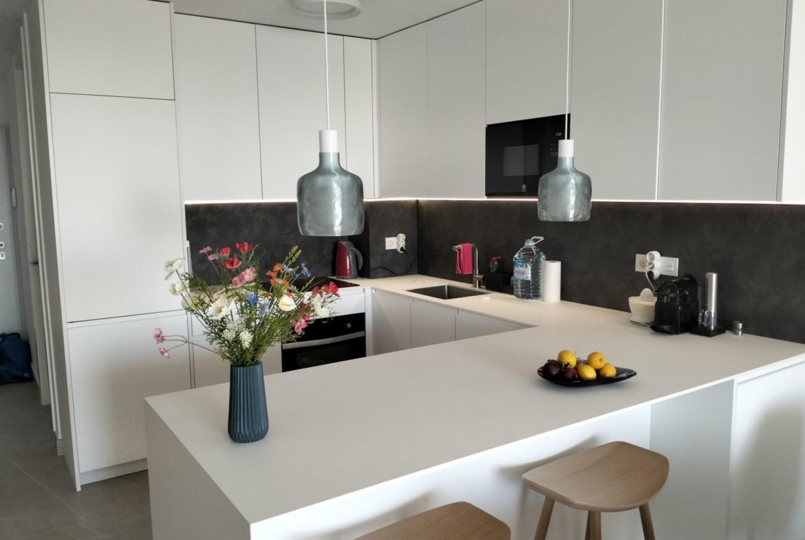 Modern white kitchen with an island, two gray pendant lamps, and a vase of colorful flowers on the counter.