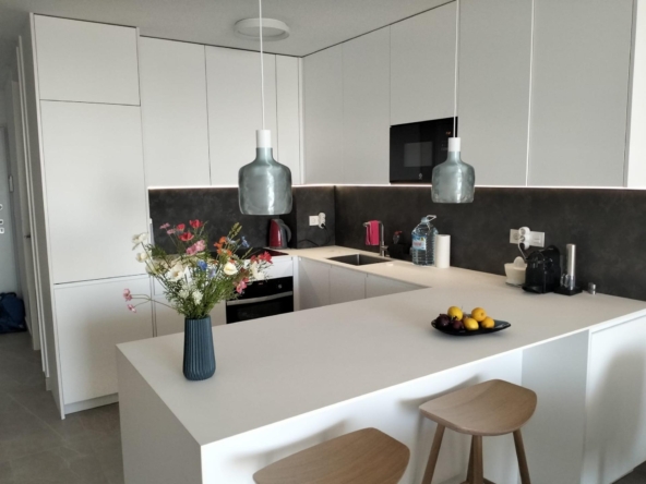 Modern white kitchen with an island, two gray pendant lamps, and a vase of colorful flowers on the counter.