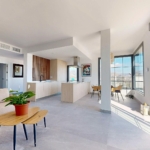 Open-plan living area with a beige sofa, wooden coffee tables, a kitchen island, and large windows with a mountain view outsideby the balcony.