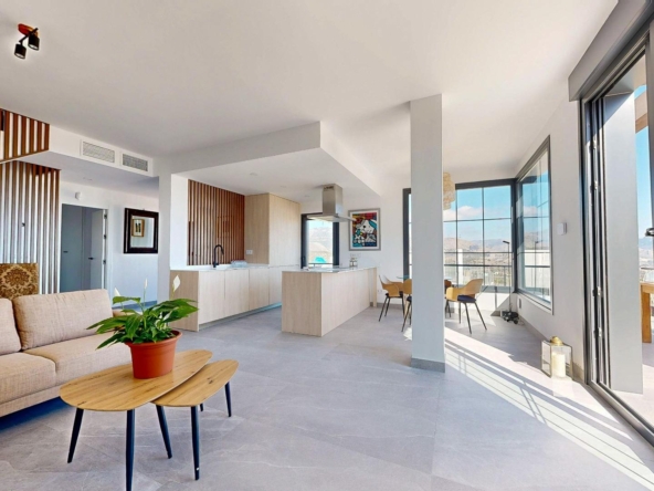 Open-plan living area with a beige sofa, wooden coffee tables, a kitchen island, and large windows with a mountain view outsideby the balcony.